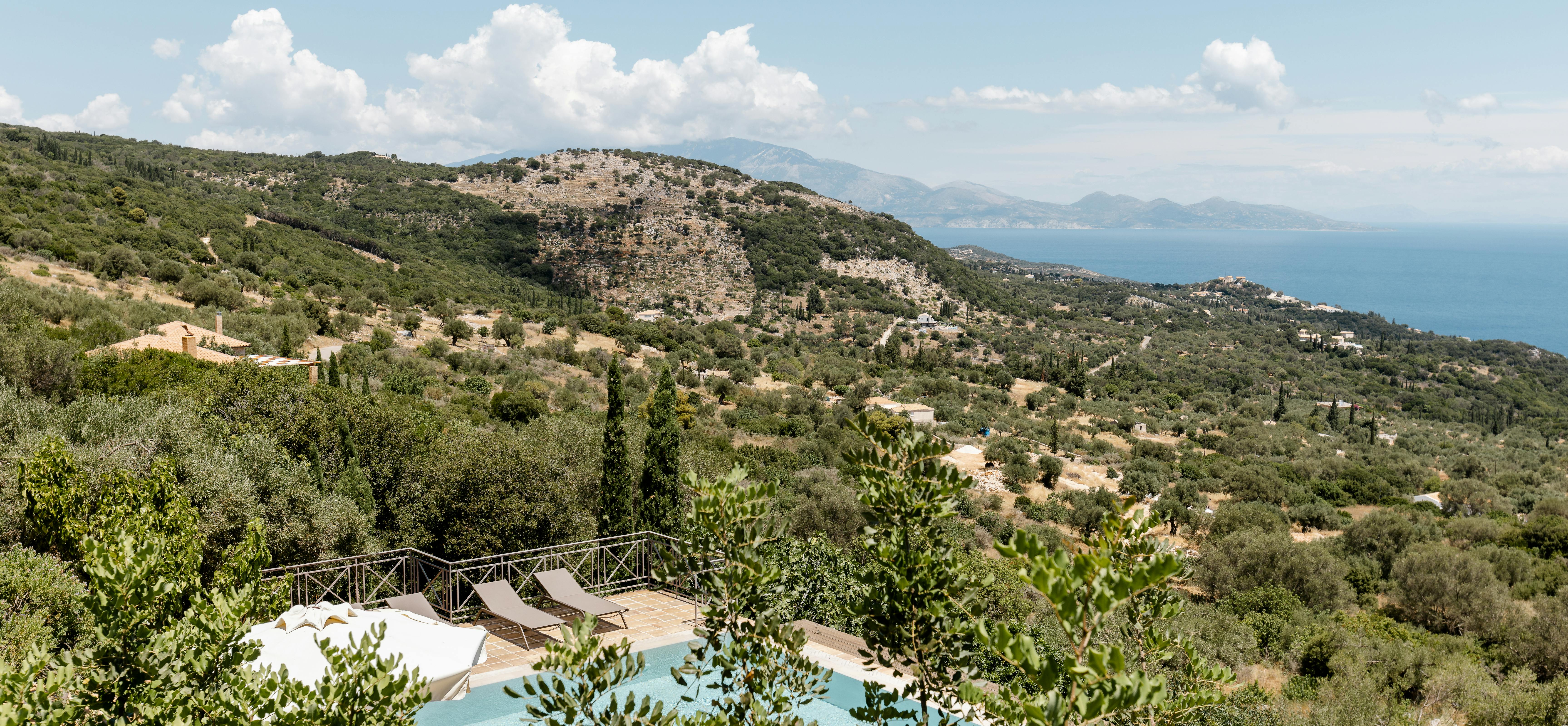 A hillside Mediterranean infinity pool with lounge chairs overlooking terraced olive groves, mountains, and the sea under a blue sky.