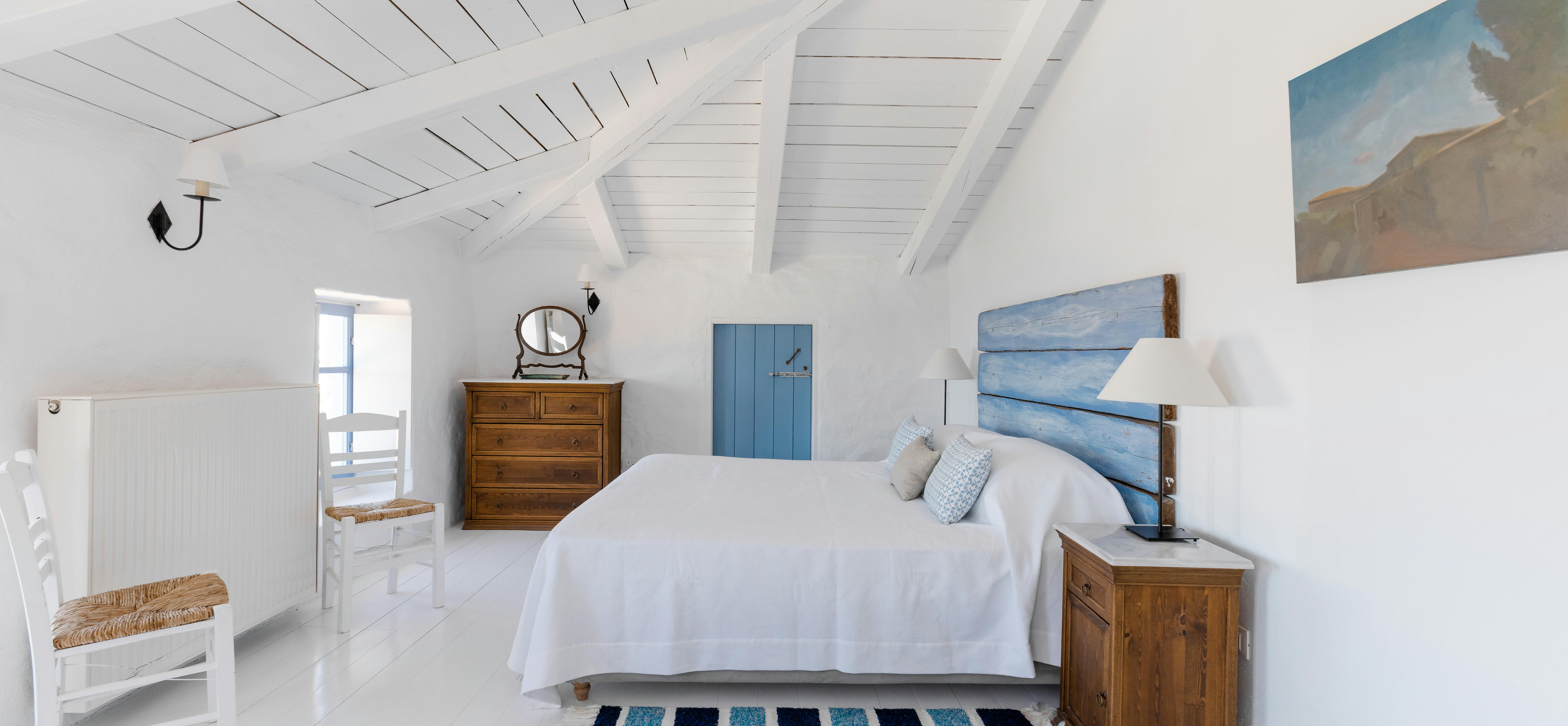 A white coastal bedroom with vaulted wood plank ceiling, distressed blue horizontal plank headboard, white linens, antique wood furniture, and blue striped rug on white painted floors.