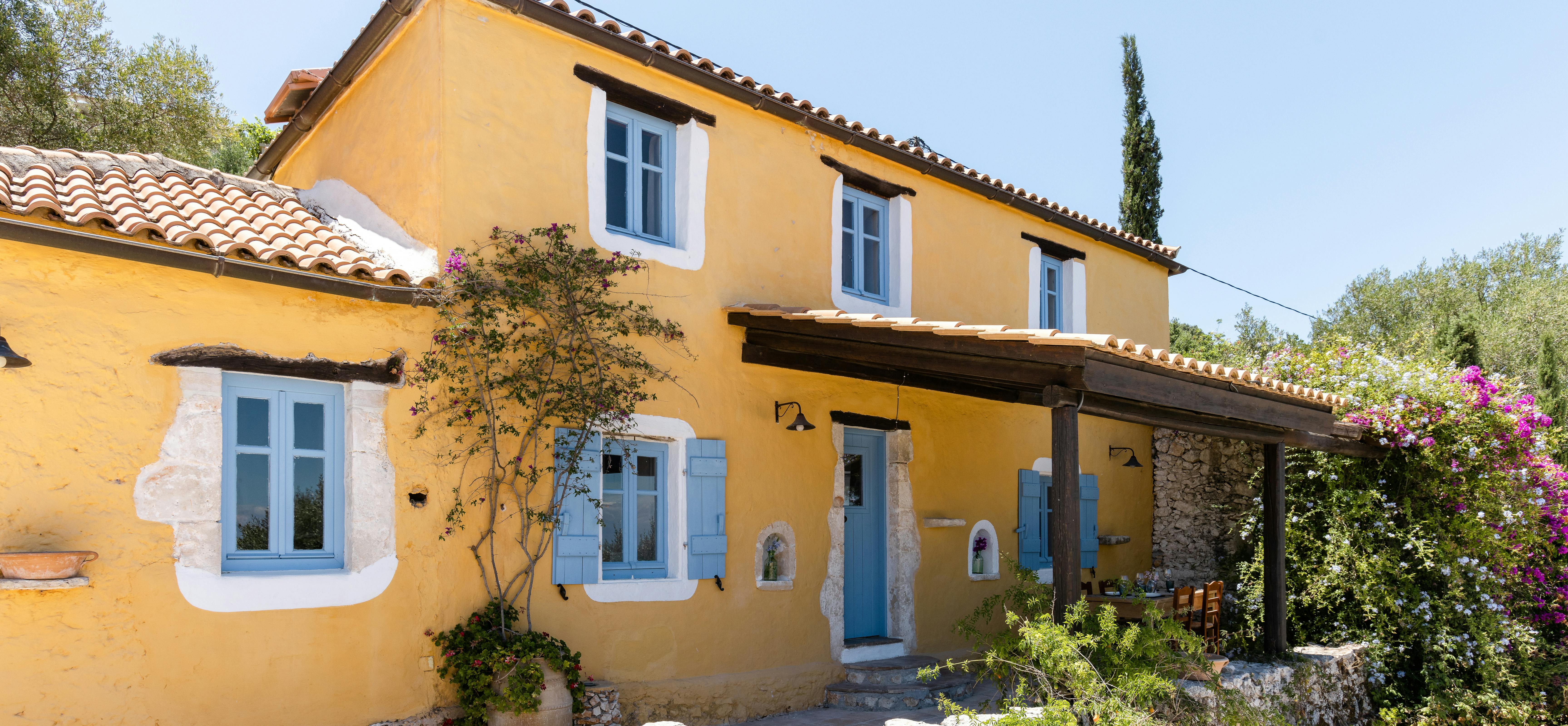 A charming ochre-yellow Mediterranean villa exterior with blue shuttered windows, terracotta tile roof, climbing vines, and lush garden with bougainvillea.