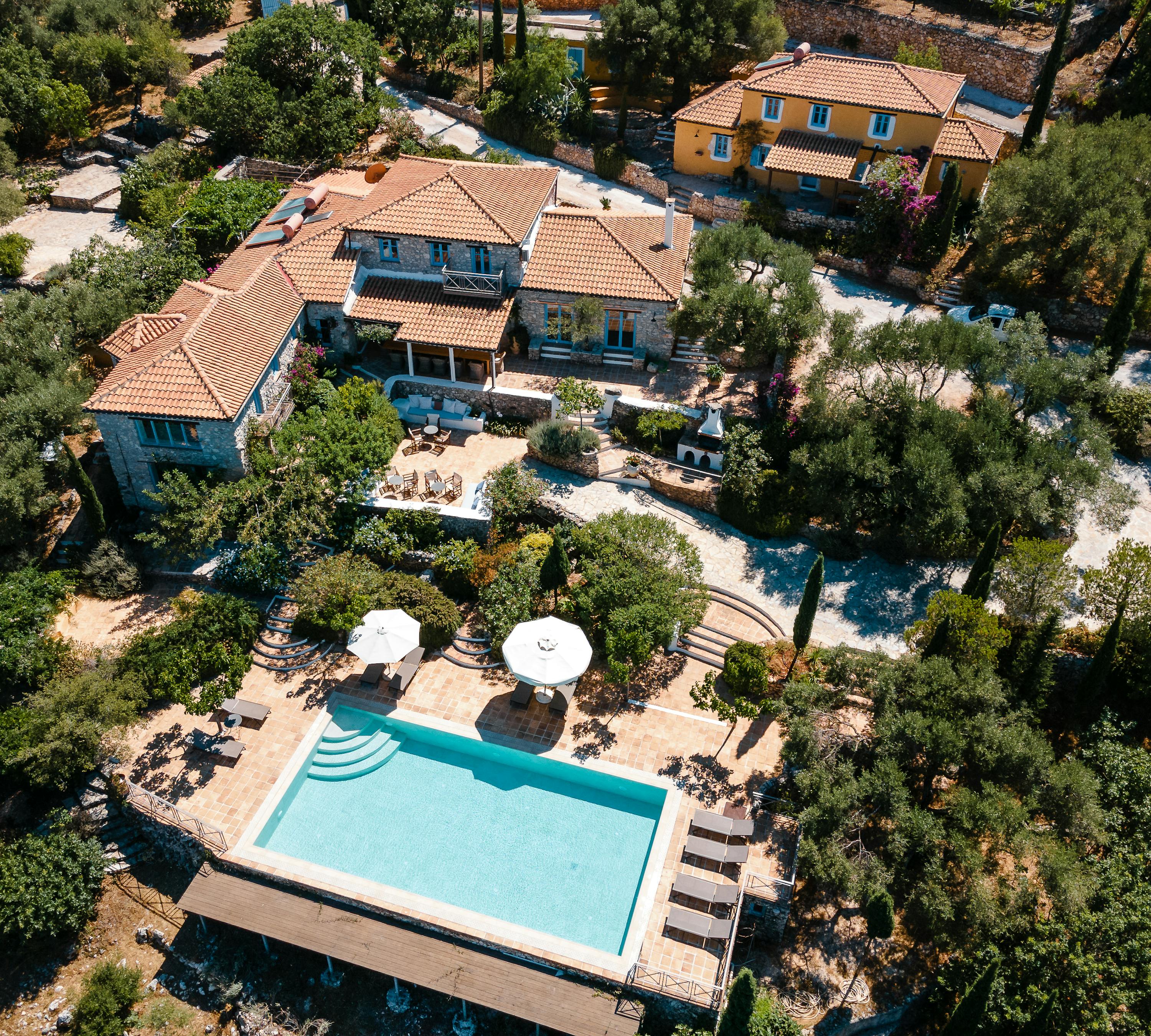 An aerial view of a Mediterranean villa estate with stone buildings, terracotta roofs, turquoise infinity pool, white umbrellas, and surrounding olive groves.