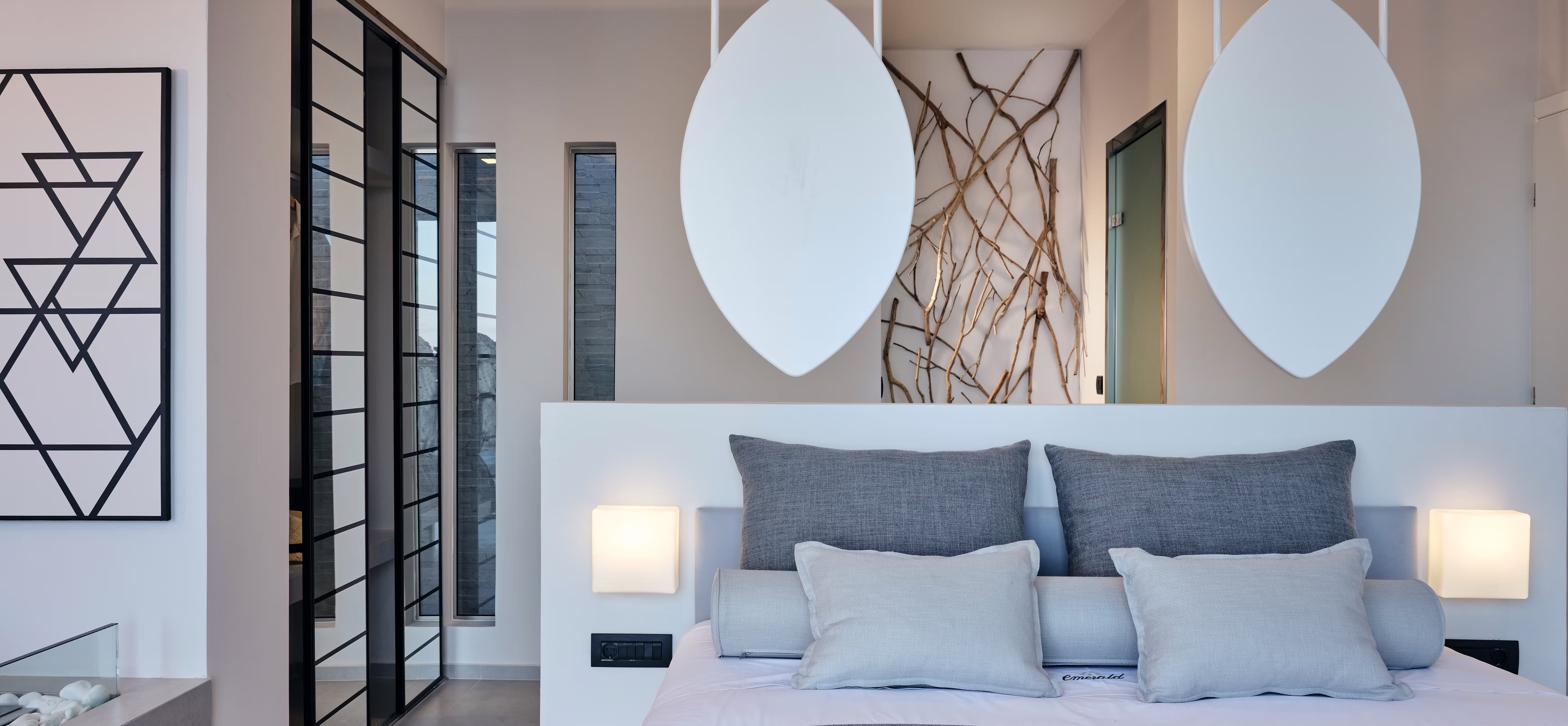 Modern bedroom featuring white platform bed with grey bedding, oversized teardrop pendant lights, and black-framed glass partitions.