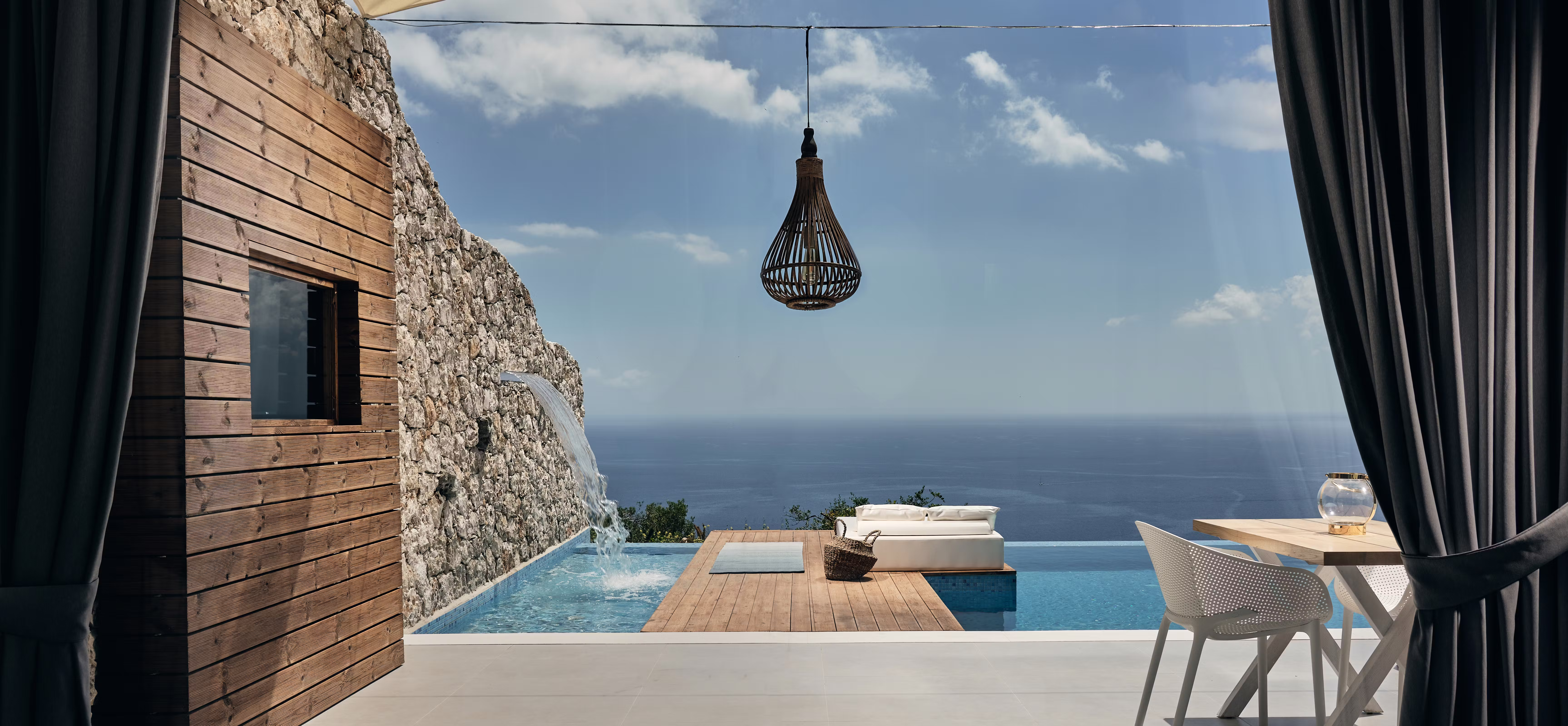 Bedroom view framing outdoor terrace with infinity pool, wooden deck, hanging lantern, and endless ocean horizon through dark curtains.