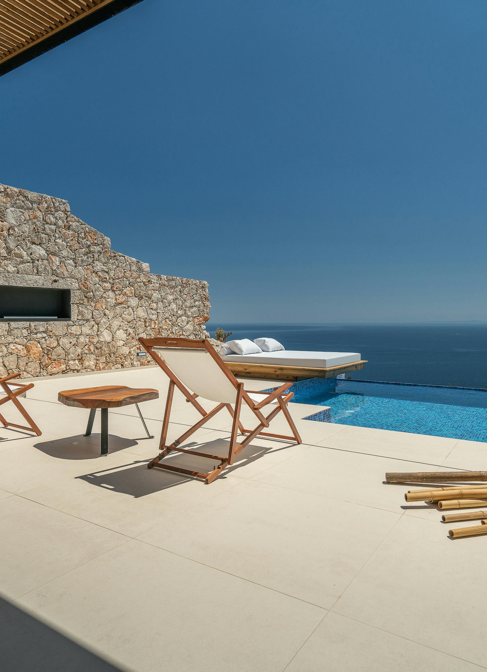 Covered terrace with wooden folding chairs and side table overlooking infinity pool, white daybed platform, and endless ocean.