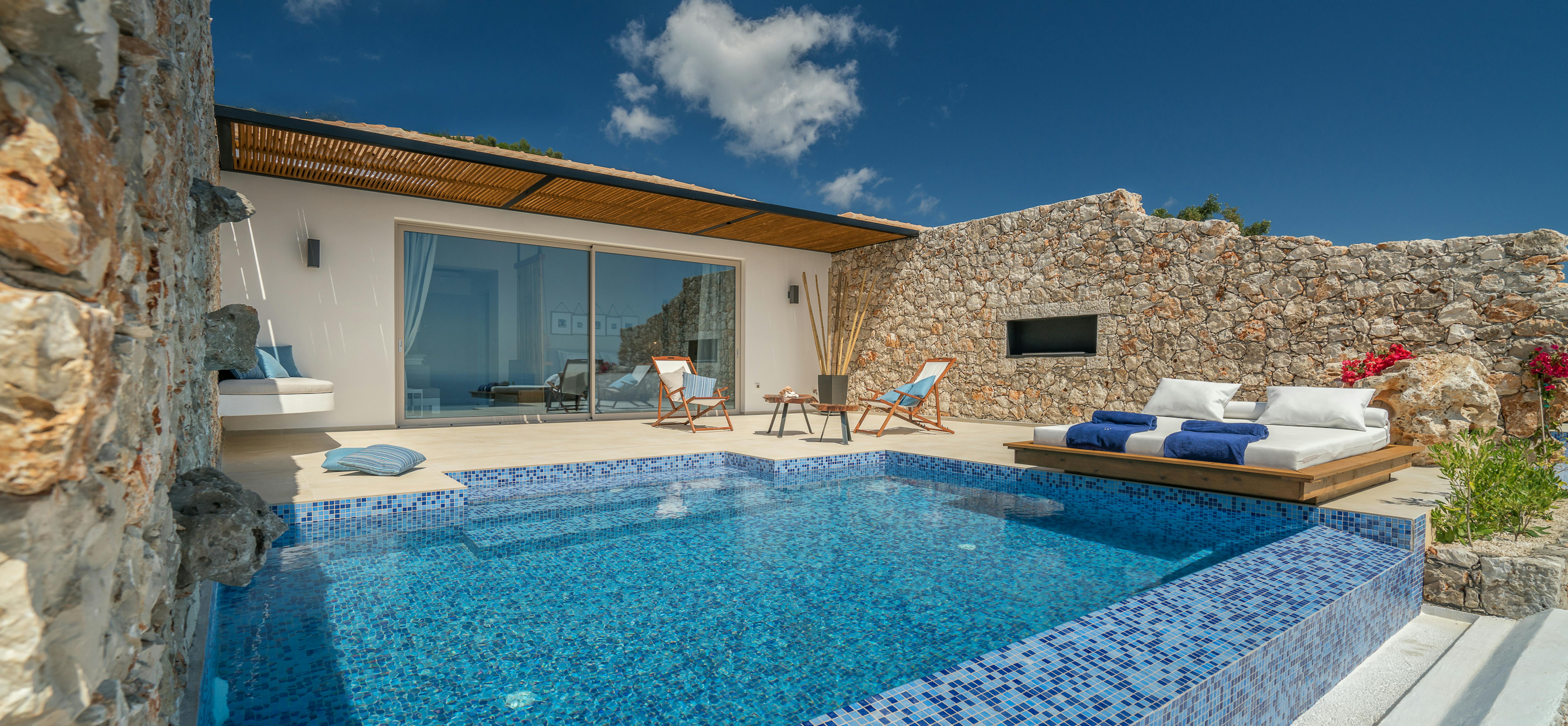 Luxury villa pool terrace with blue mosaic-tiled infinity pool, wooden platform daybed, stone walls, and modern pavilion with slatted ceiling.