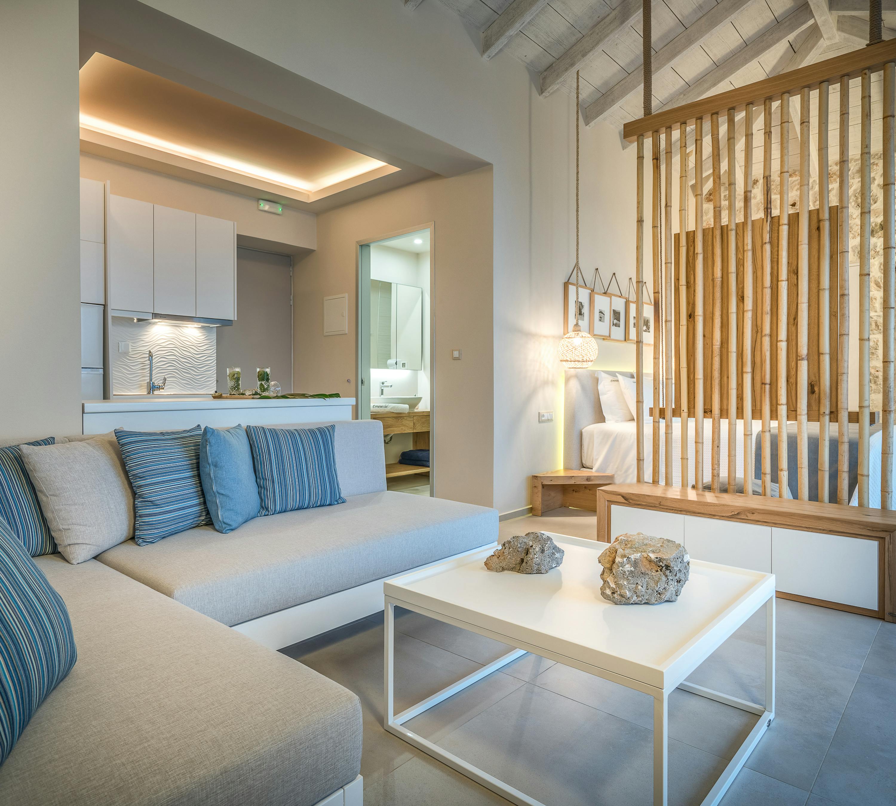 Open-plan living space with blue striped sectional sofa, vertical wood slat divider, exposed beam ceiling, and kitchen with ocean view.