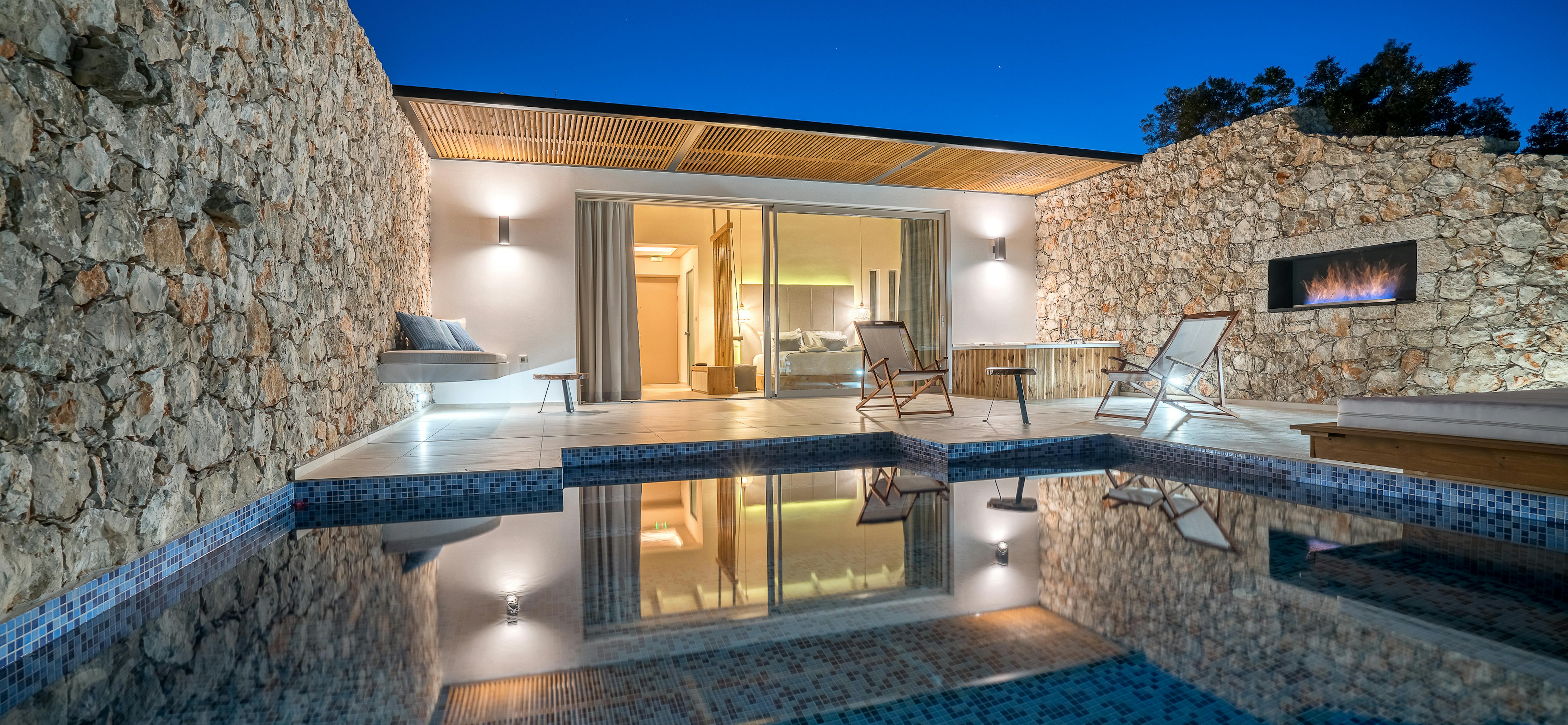 Evening reflection of illuminated villa in still infinity pool surrounded by ancient stone walls and outdoor furniture at dusk.