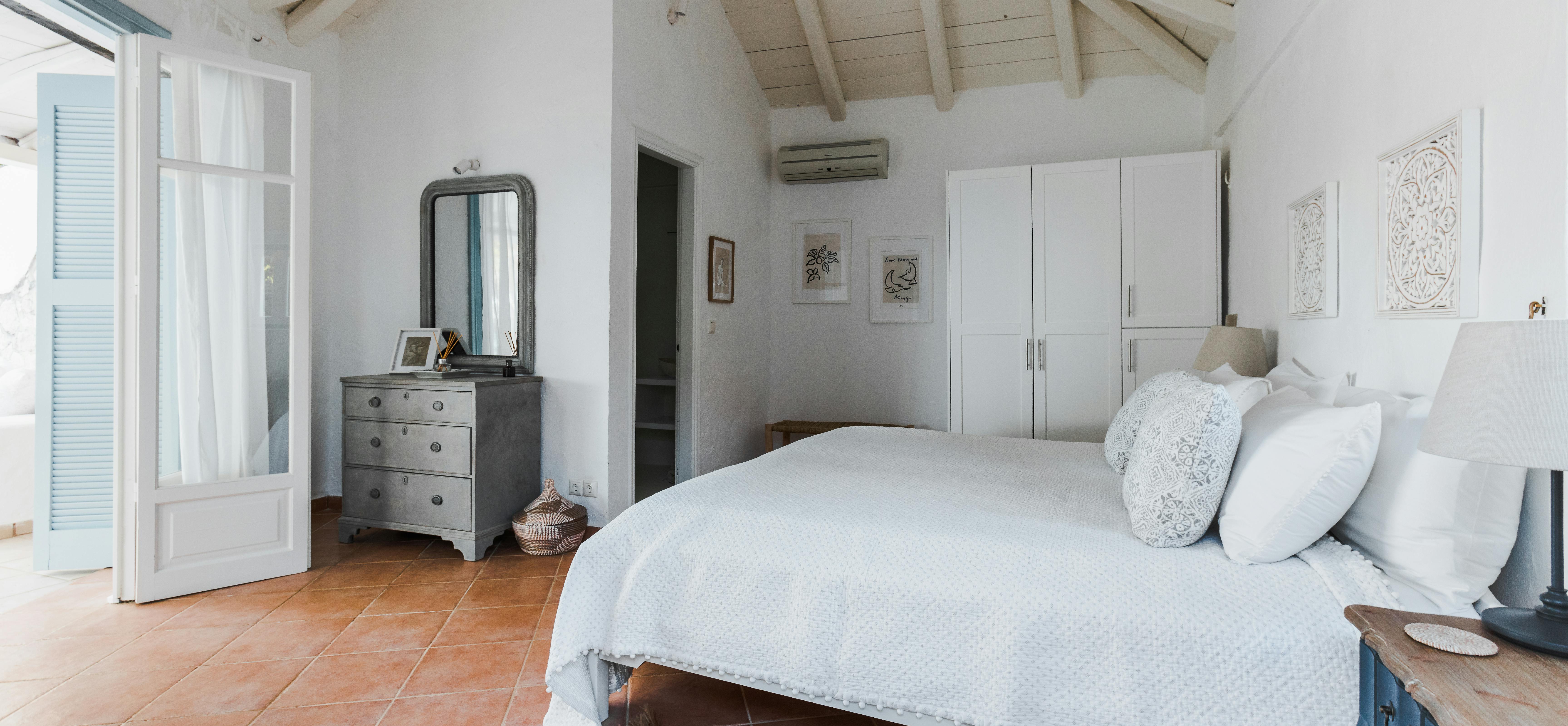 White coastal bedroom with vaulted ceiling, vintage gray dresser, arched mirror, white bedding, and terracotta tile flooring.
