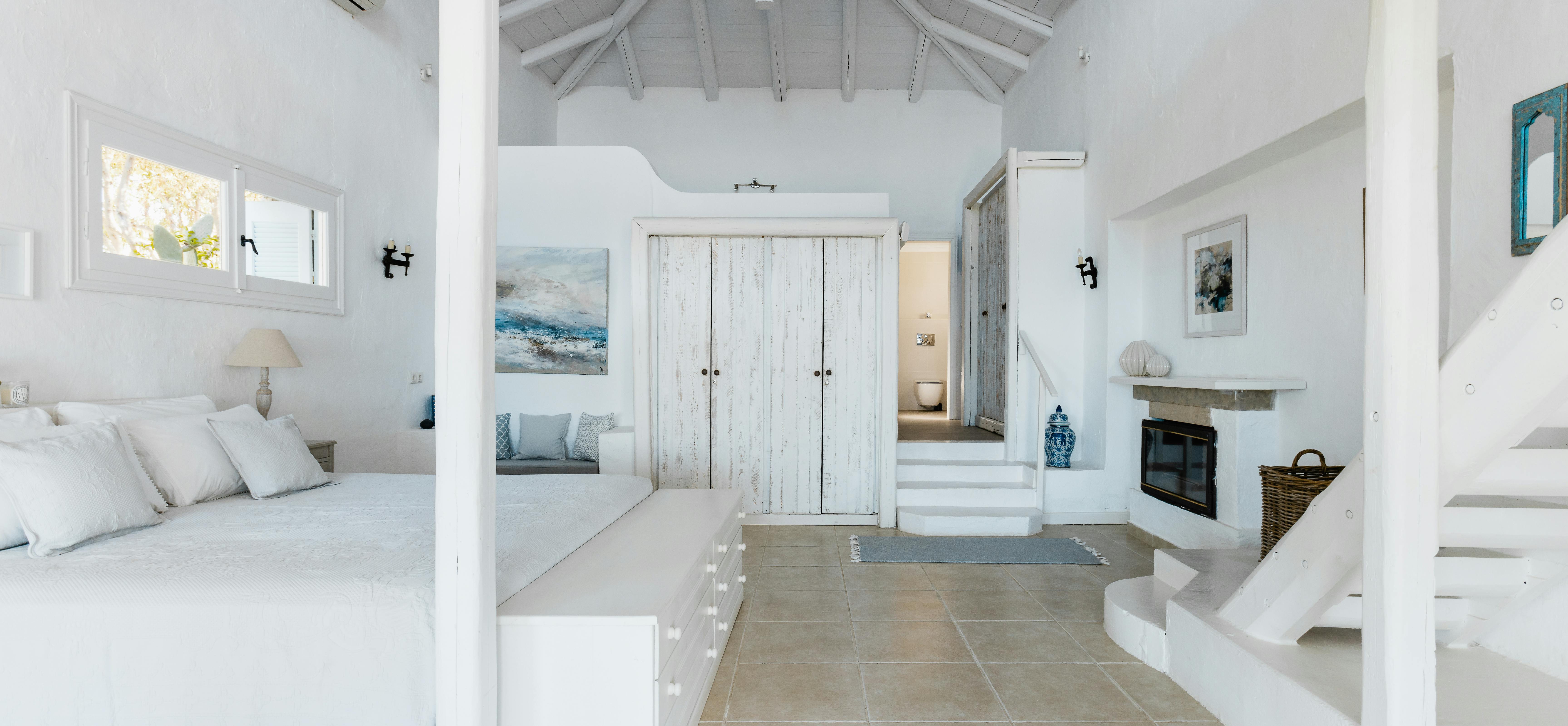 Airy white bedroom suite with vaulted wood ceiling, whitewashed wardrobe, built-in window seat with coastal artwork, and terracotta tile floors leading to sitting area.