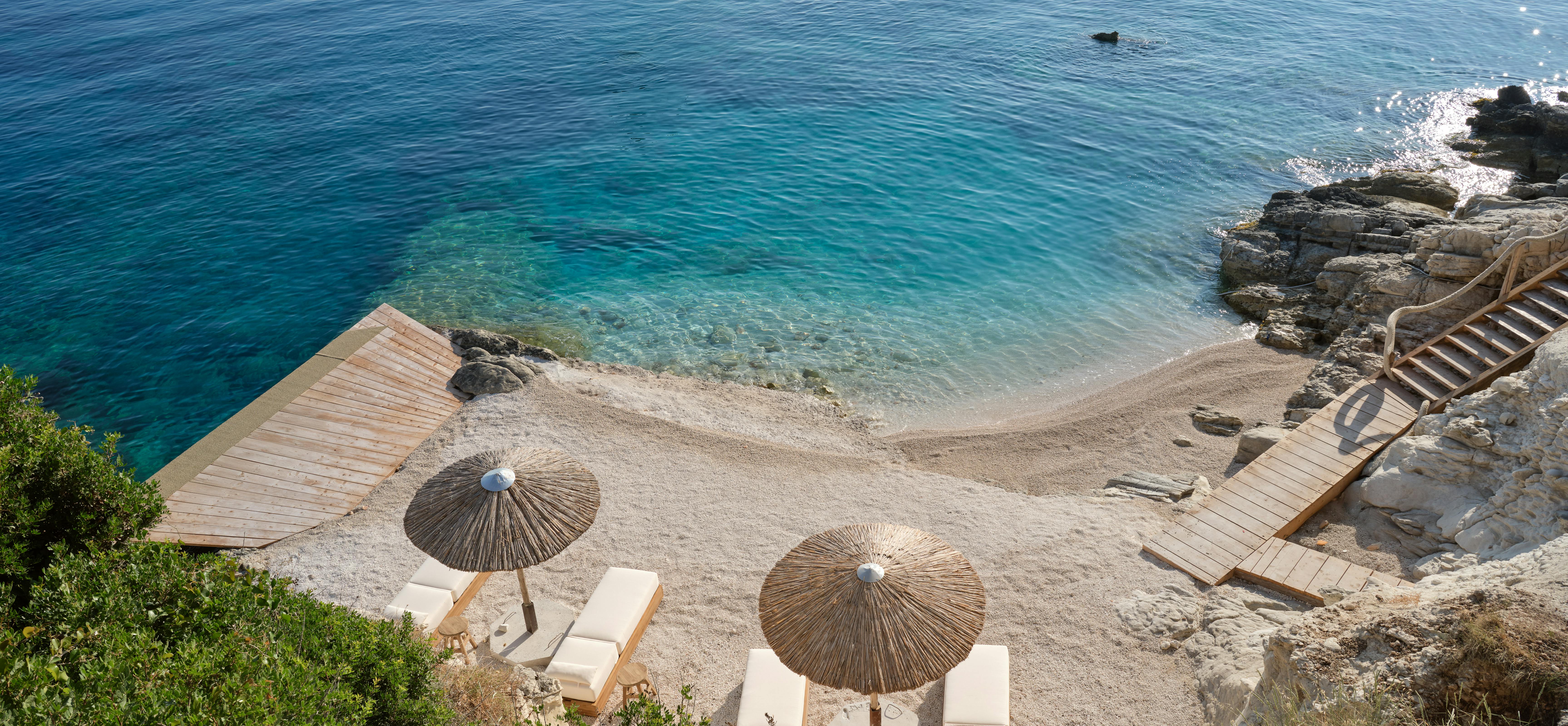 An aerial view reveals a private crescent beach with crystalline turquoise waters lapping against pale sand, where wooden boardwalk paths connect the shore to teak loungers shaded by natural thatched umbrellas positioned on the intimate cove.