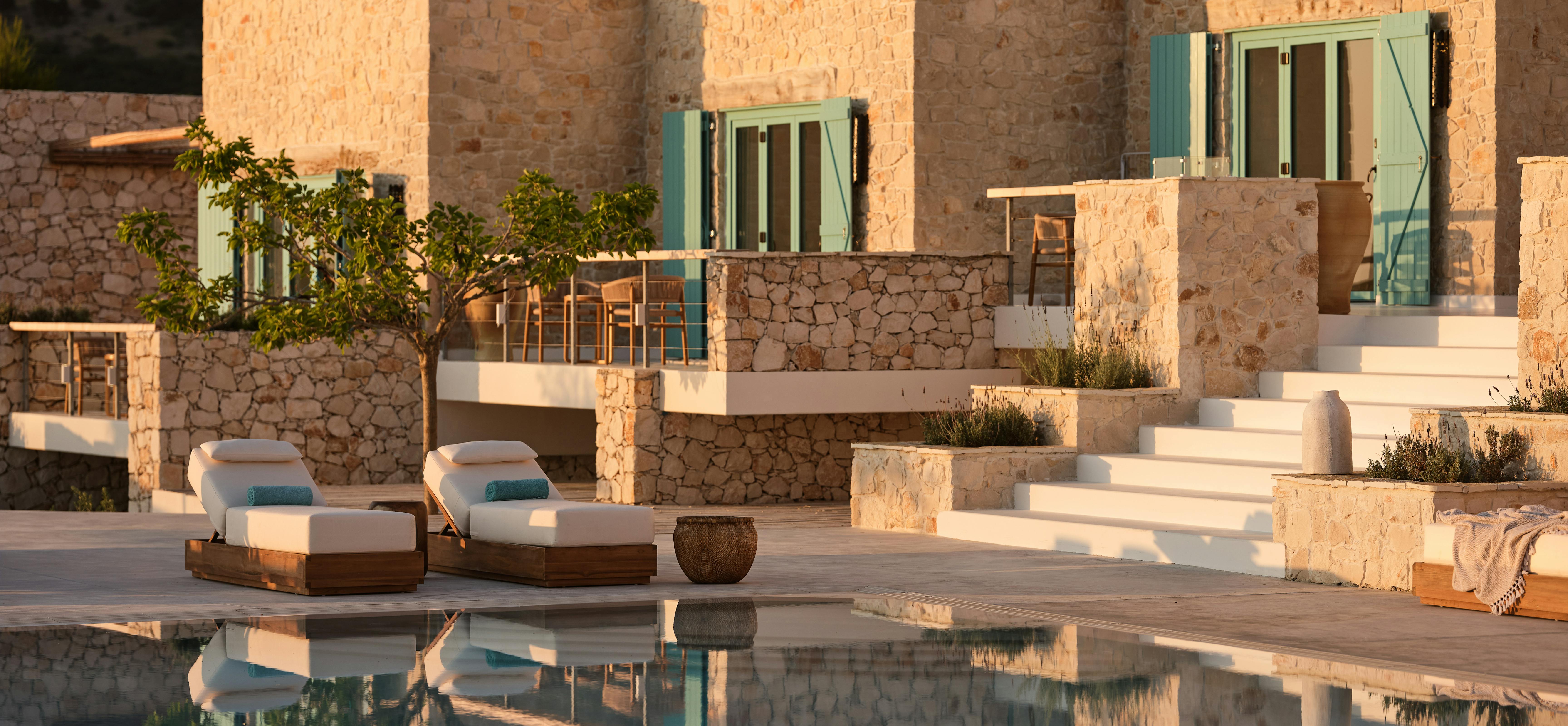 The mirror-like infinity pool captures pristine reflections of a multi-level Mediterranean villa built from local limestone with soft mint-green shutters, white terraced steps adorned with planters, and teak furniture on the pool deck as gentle light washes over the scene.