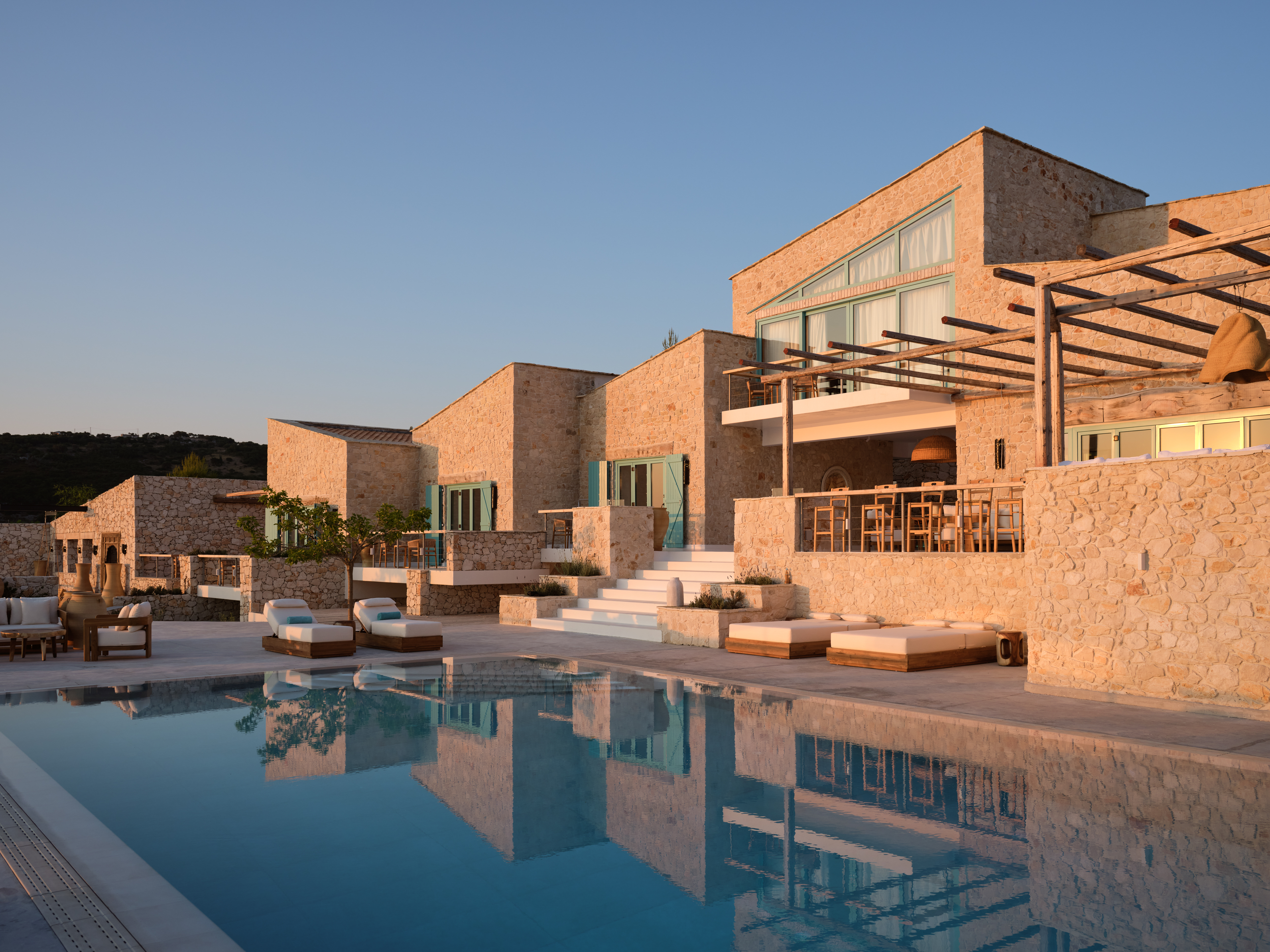 The infinity pool's turquoise water creates stunning reflections of the multi-story limestone villa complex bathed in golden evening light, showcasing the terraced architecture with mint-green shuttered windows, wooden pergolas, white stone steps, and scattered teak loungers.
