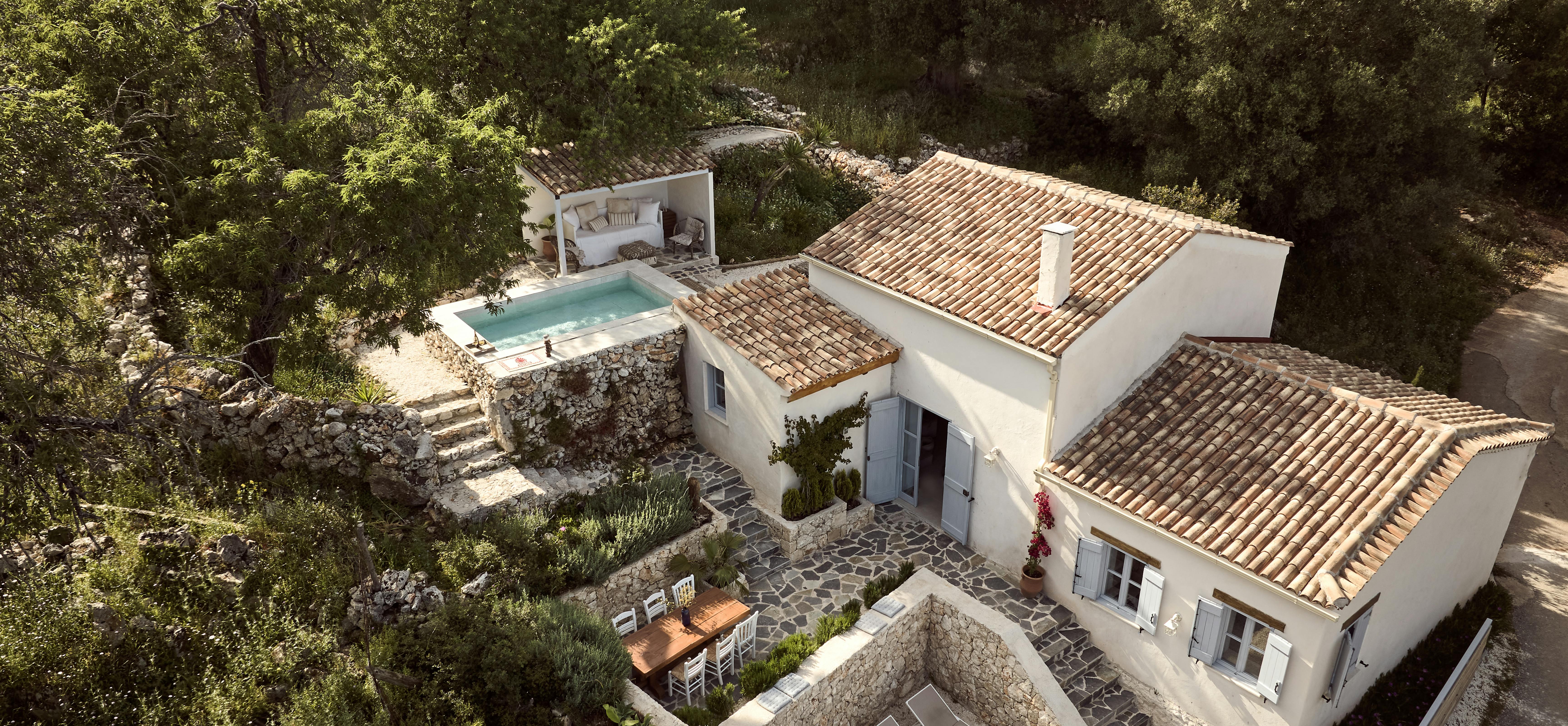 An aerial view of a white Mediterranean villa with terracotta tile roofing nestled in lush greenery, featuring a pool with a covered lounge area and stone terraced outdoor spaces.