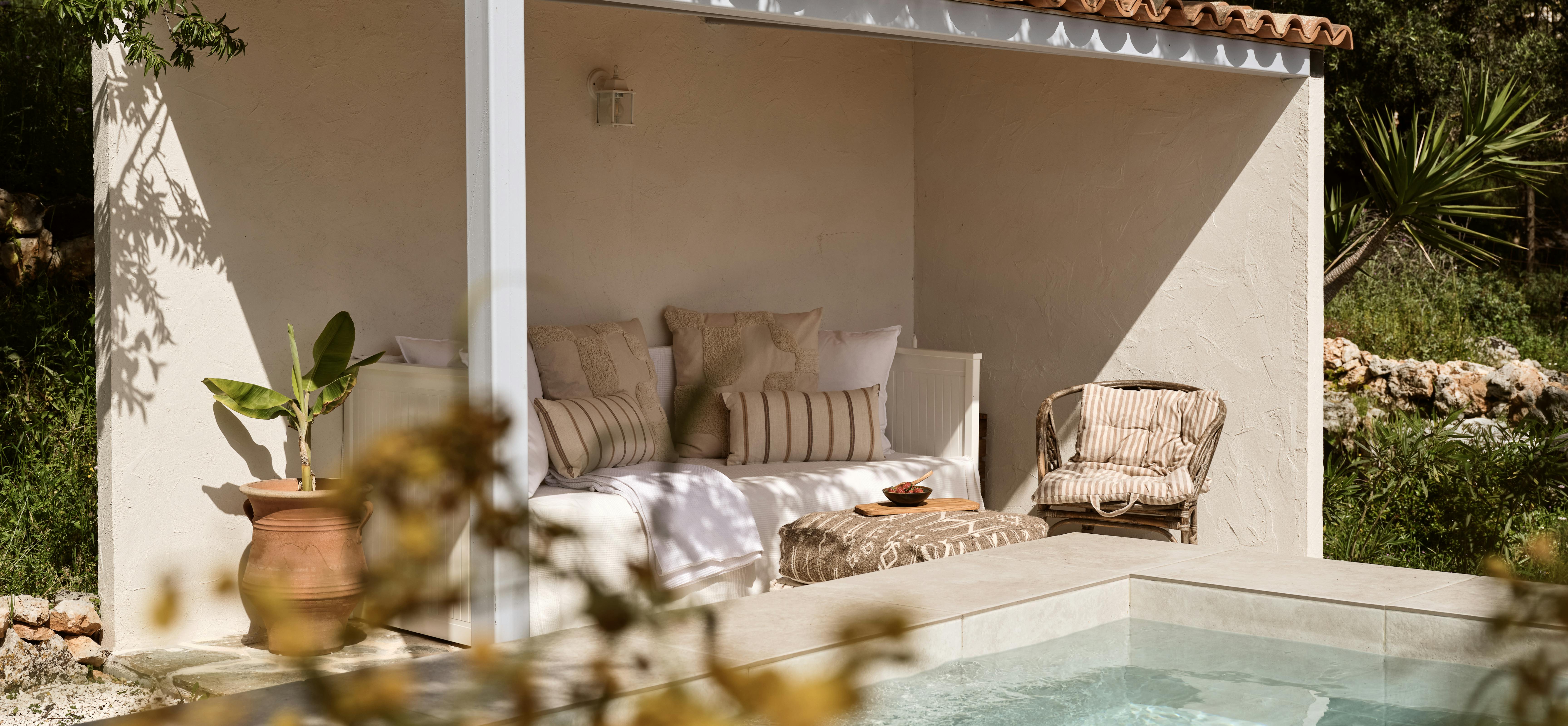 A covered outdoor daybed with white cushions and striped pillows beside a pool, set within a white-walled pavilion with terracotta tile roof surrounded by Mediterranean gardens.
