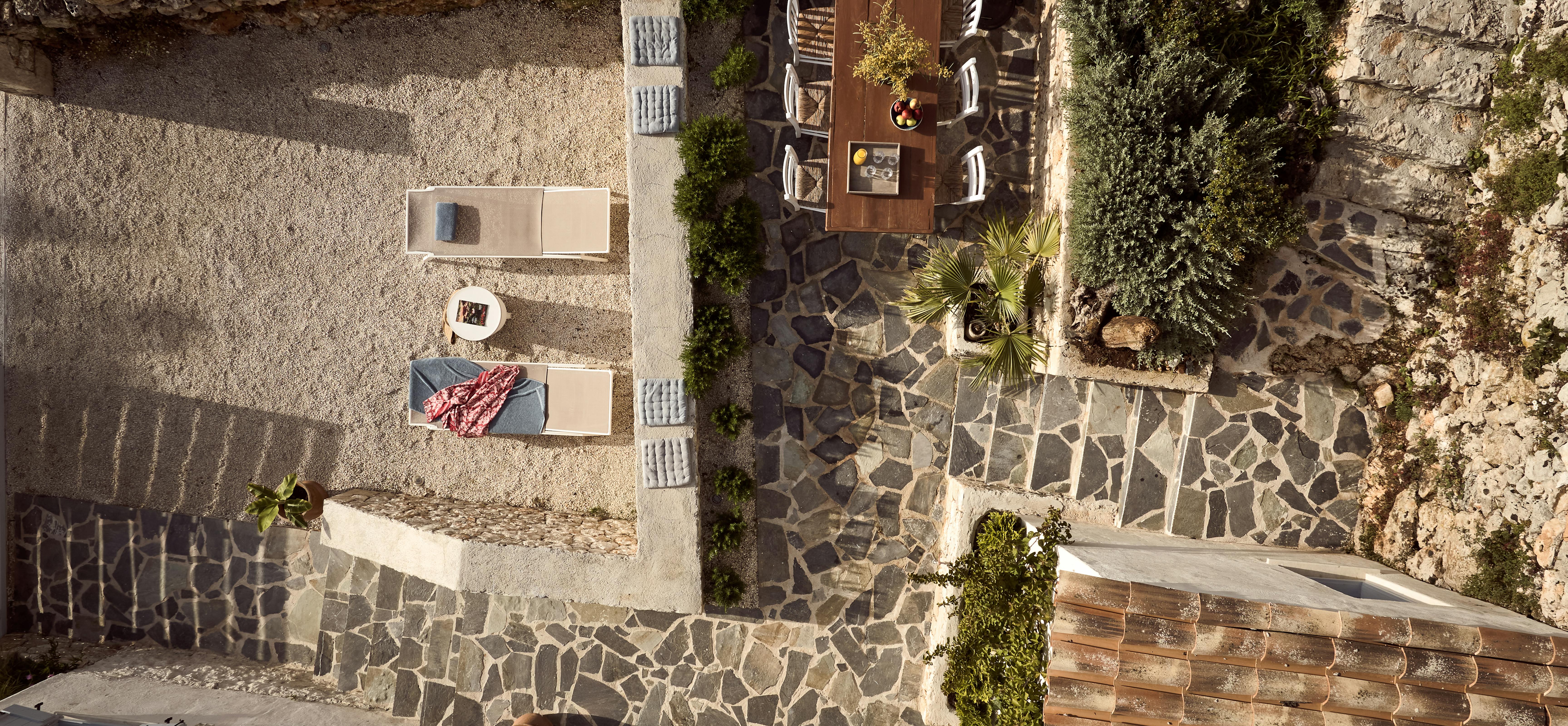 An aerial view of multi-level stone terraces with mosaic stonework, showing an outdoor dining area, sun loungers, and various Mediterranean landscaping elements.