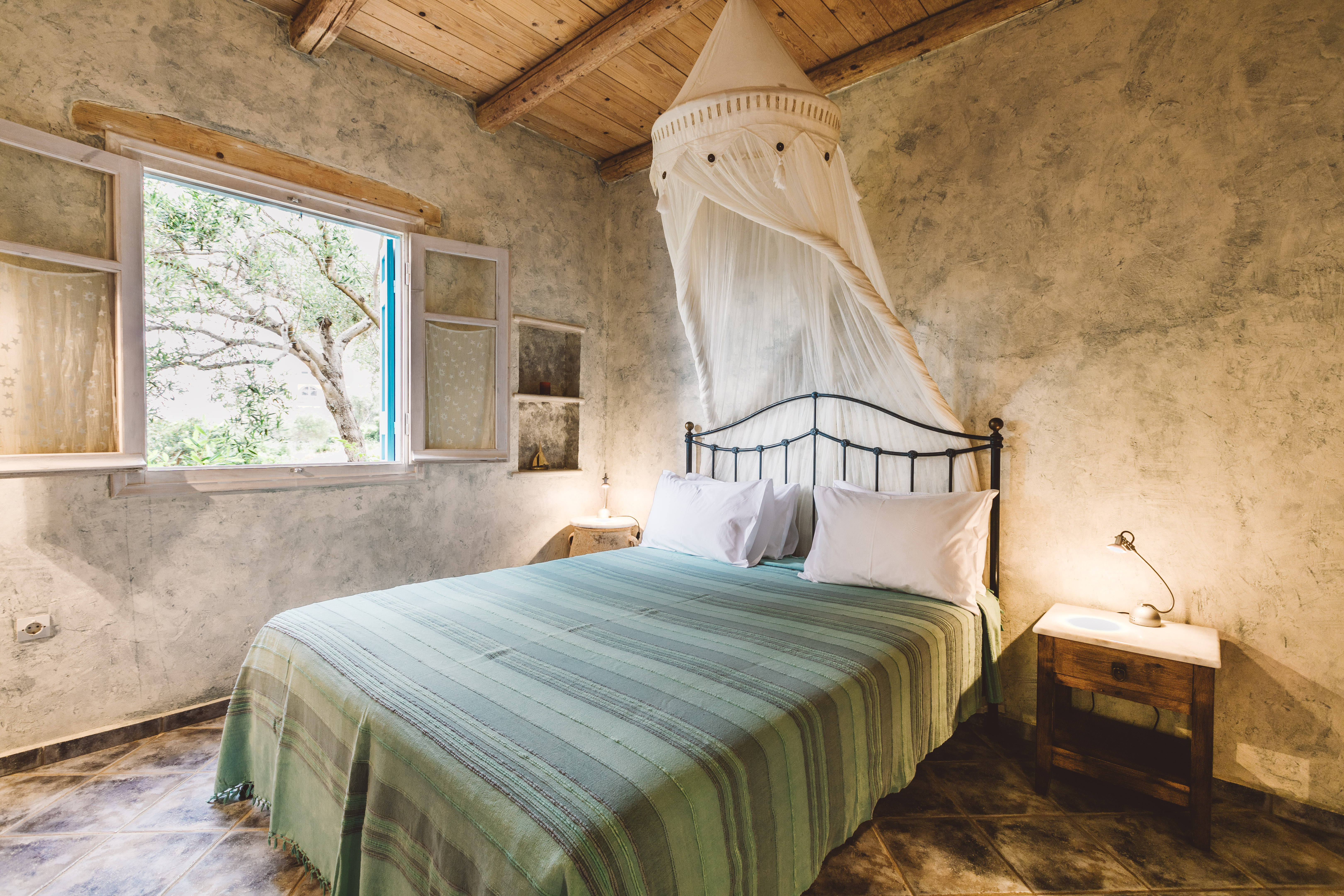A rustic bedroom with weathered plaster walls, exposed wooden ceiling beams, black iron bed with green striped bedspread, white mosquito netting canopy, and window overlooking olive trees.