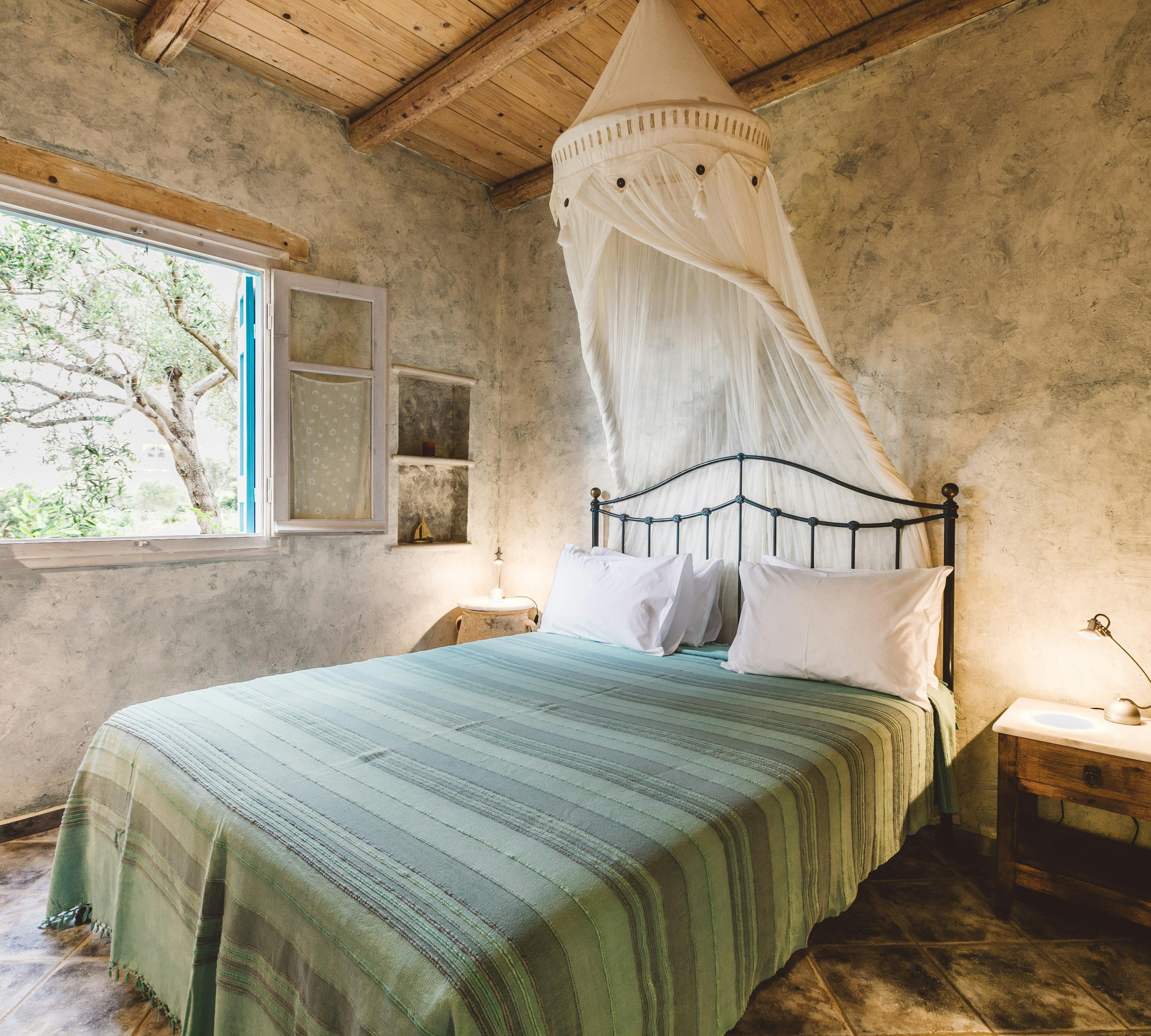 A rustic bedroom with weathered plaster walls, exposed wooden ceiling beams, black iron bed with green striped bedspread, white mosquito netting canopy, and window overlooking olive trees.