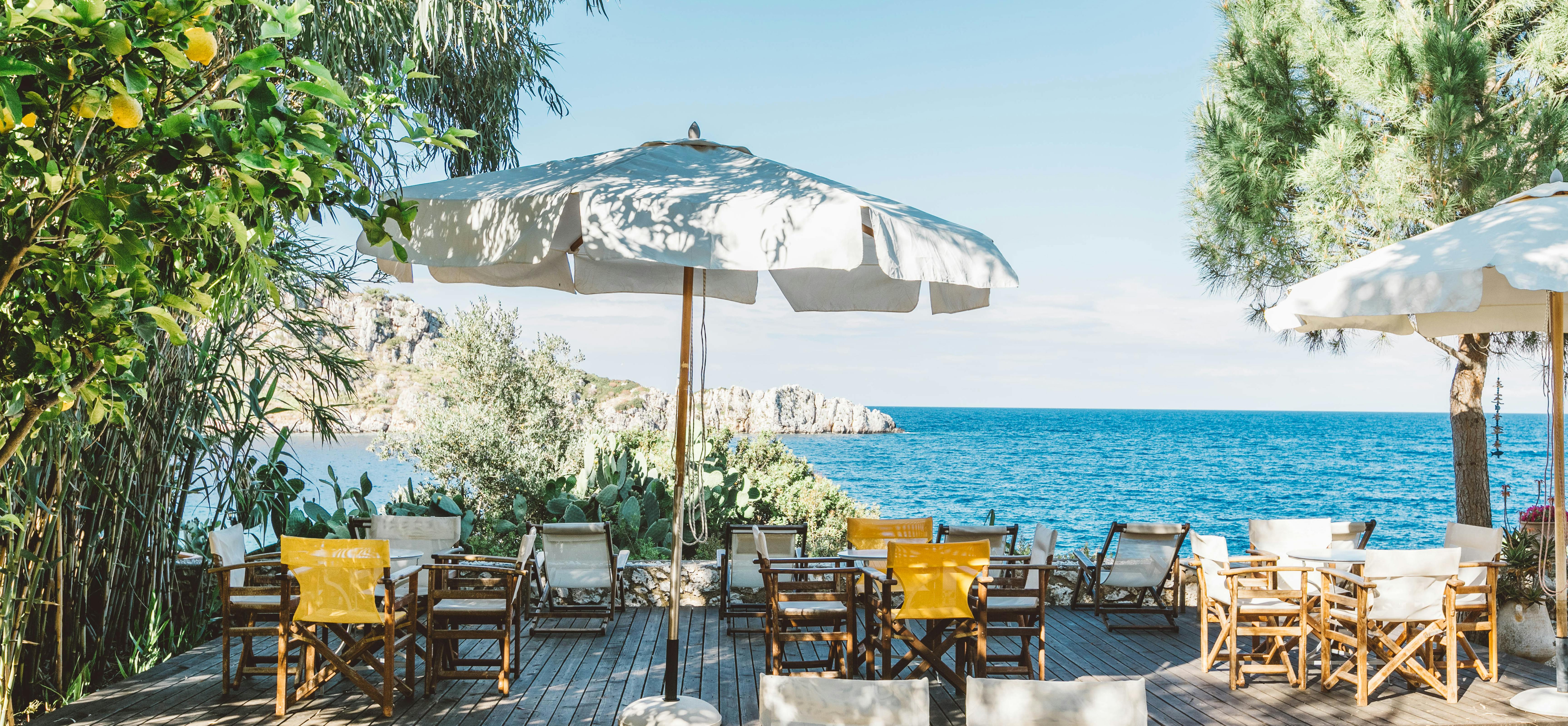 A beachfront wooden deck with canvas director's chairs, white and yellow cushions, striped umbrellas, citrus trees, and blue Mediterranean waters visible beyond.