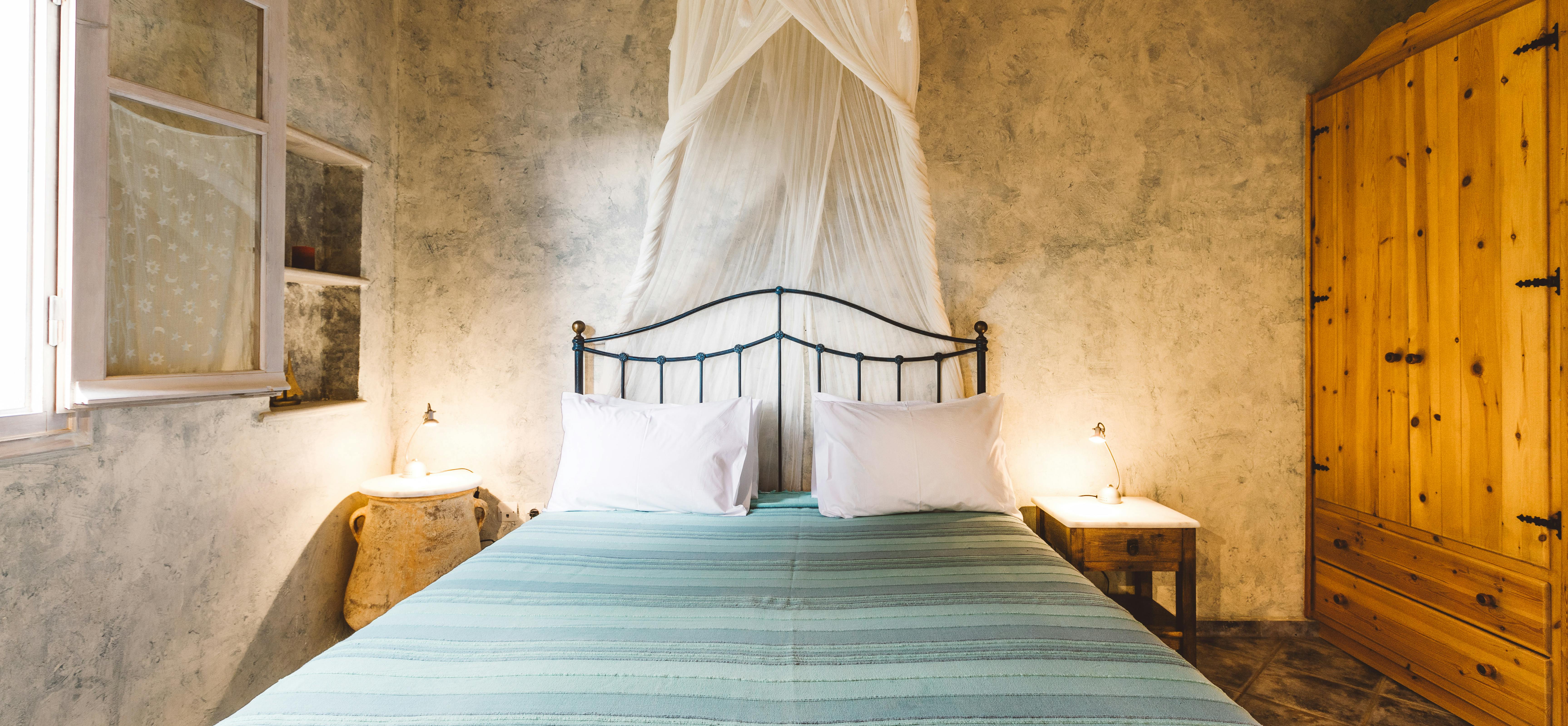 A rustic bedroom with textured plaster walls, exposed wooden ceiling, black iron bed with turquoise and green striped bedspread, white mosquito netting, wooden wardrobe, and windows with turquoise trim.