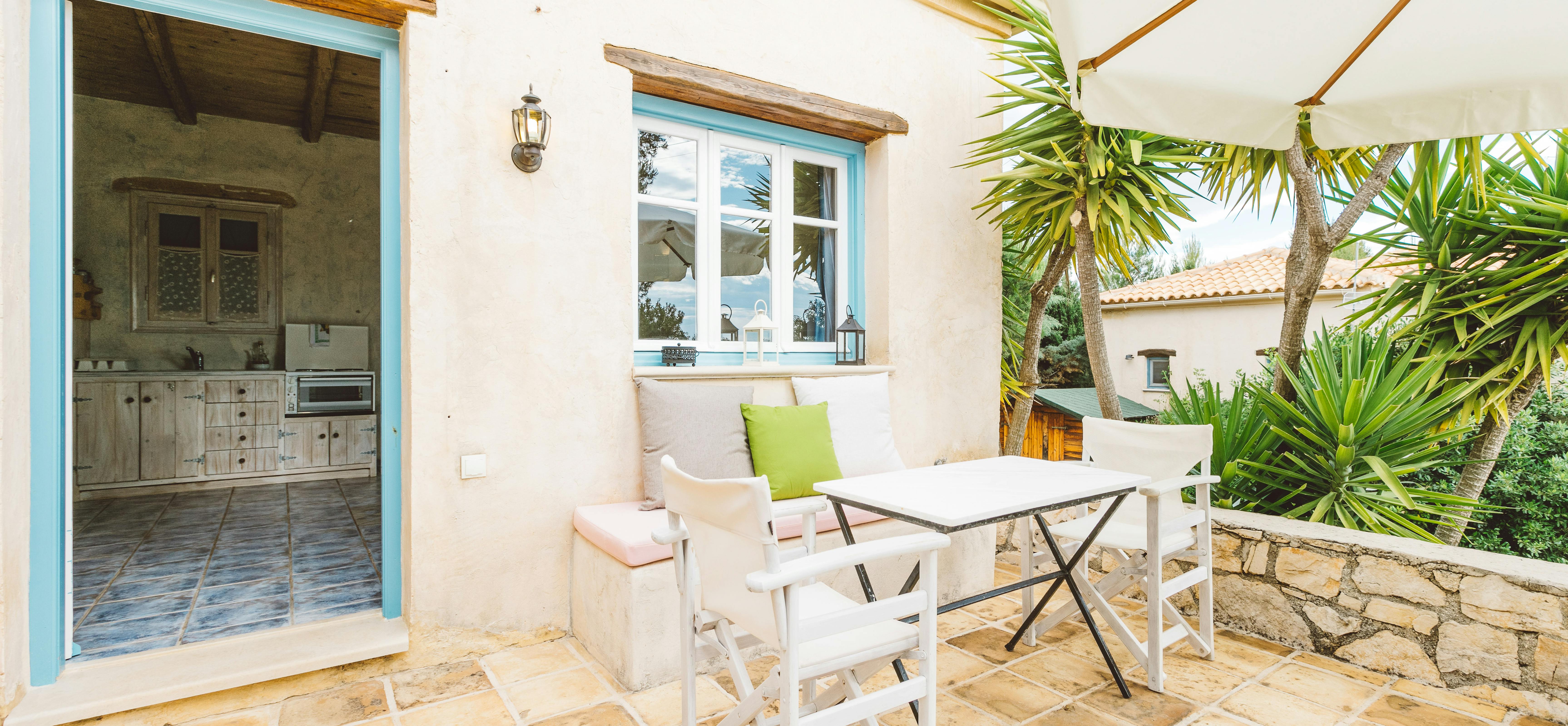 A Mediterranean terrace with white walls, turquoise-trimmed doorway and window, built-in bench seating, white folding chairs, small table under cream umbrella, palm trees, and stone paving.