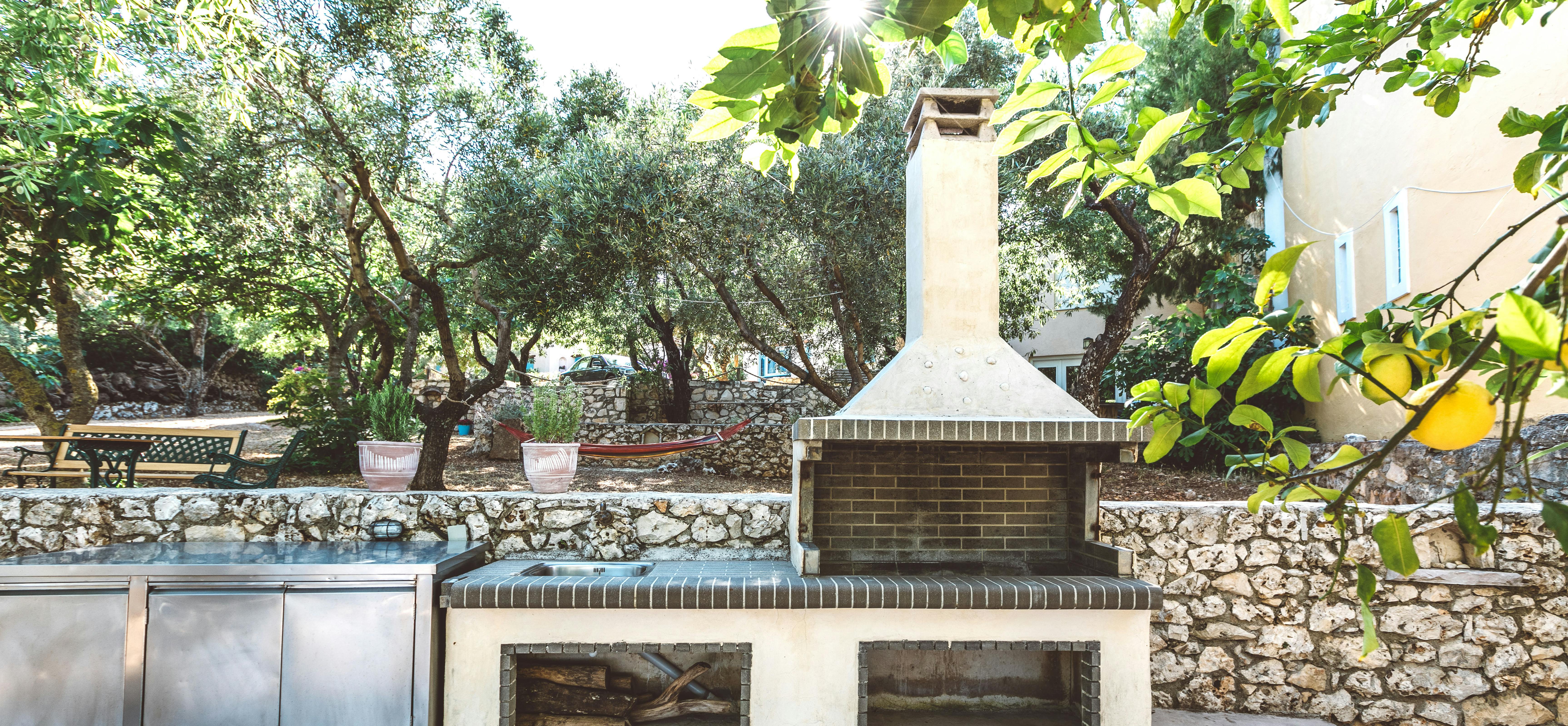 An outdoor kitchen area beneath lemon trees with white stucco barbecue and chimney, stainless steel counters, stone walls, olive grove, and dappled sunlight filtering through foliage.