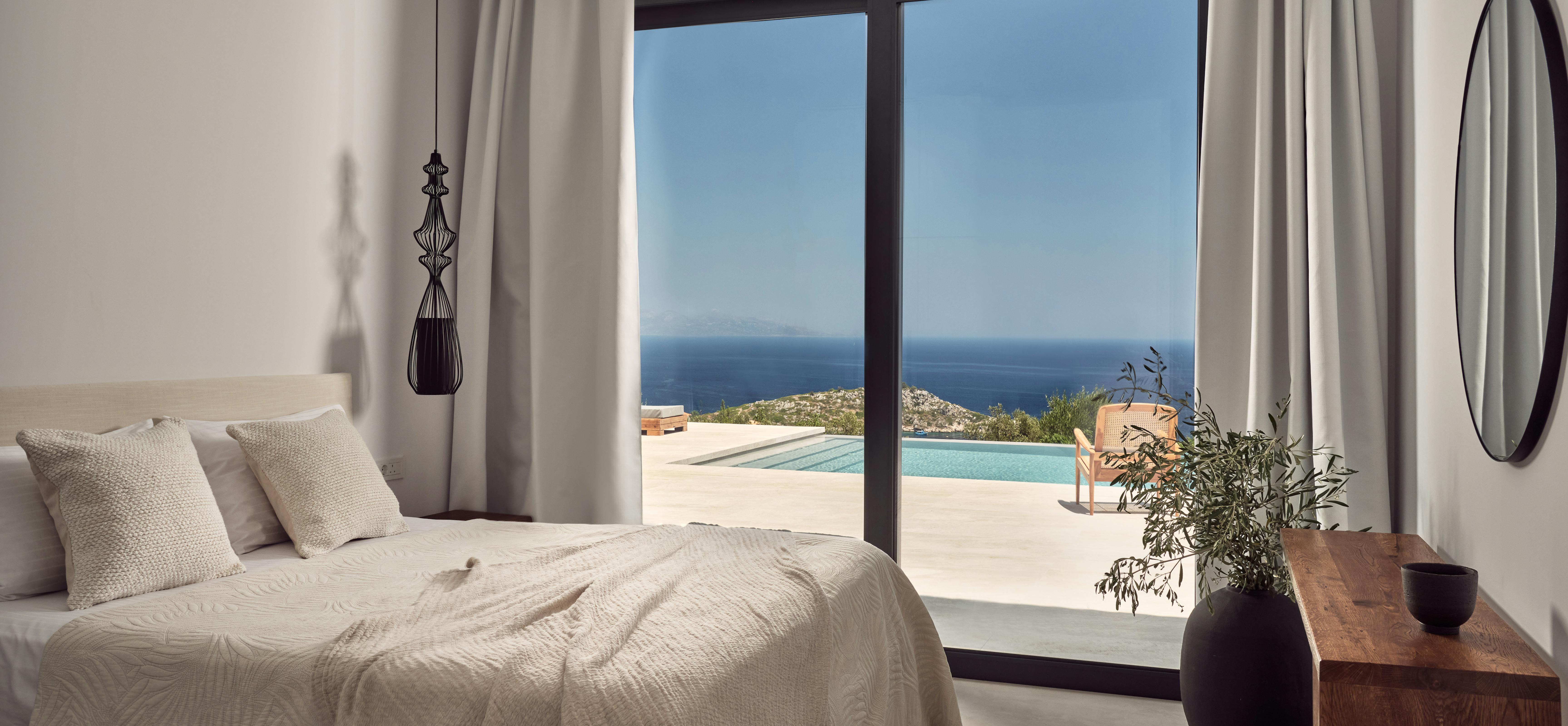 A minimalist white bedroom with floor-to-ceiling glass doors opening to an infinity pool terrace with panoramic Mediterranean Sea views, featuring a sculptural black pendant light and natural wood furnishings.