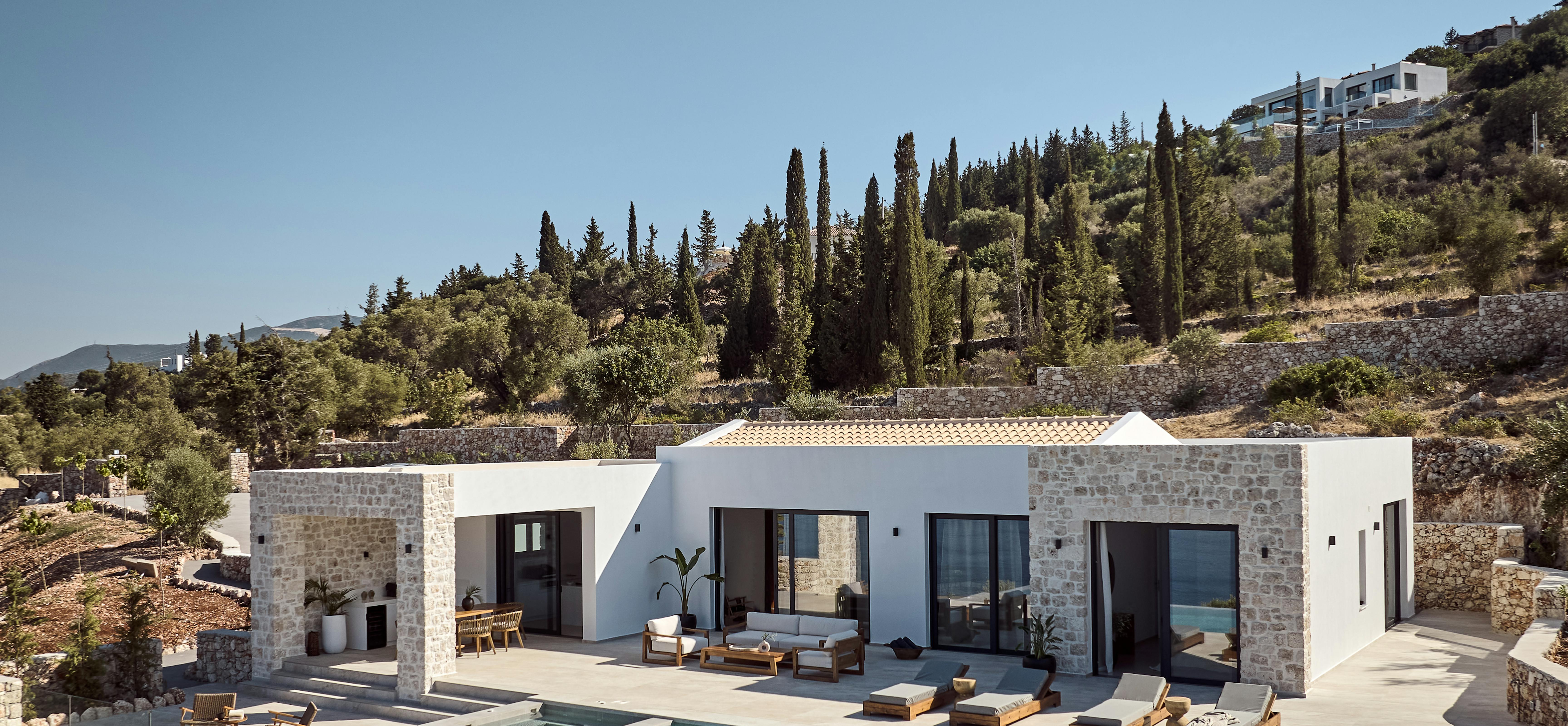 A modern hillside Mediterranean villa with white stucco and stone facades, featuring a large infinity pool surrounded by wooden sun loungers, terraced landscaping with cypress trees and olive groves climbing the slope behind.