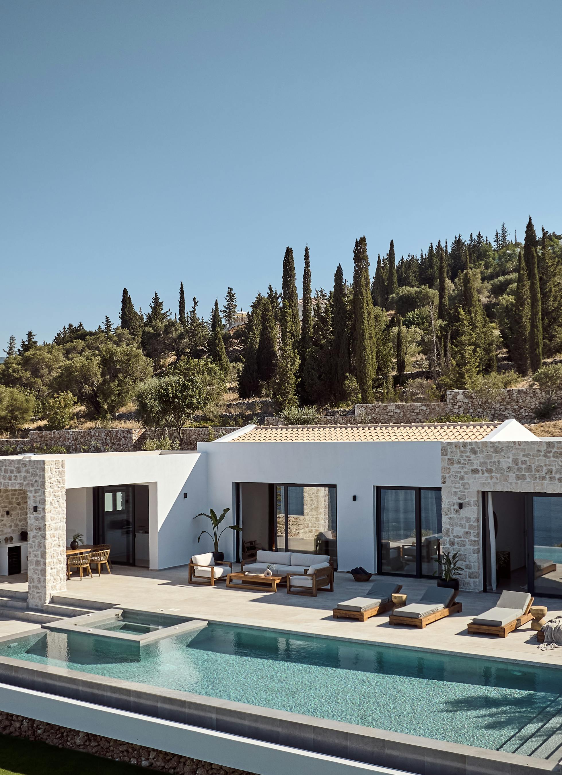 A modern hillside Mediterranean villa with white stucco and stone facades, featuring a large infinity pool surrounded by wooden sun loungers, terraced landscaping with cypress trees and olive groves climbing the slope behind.