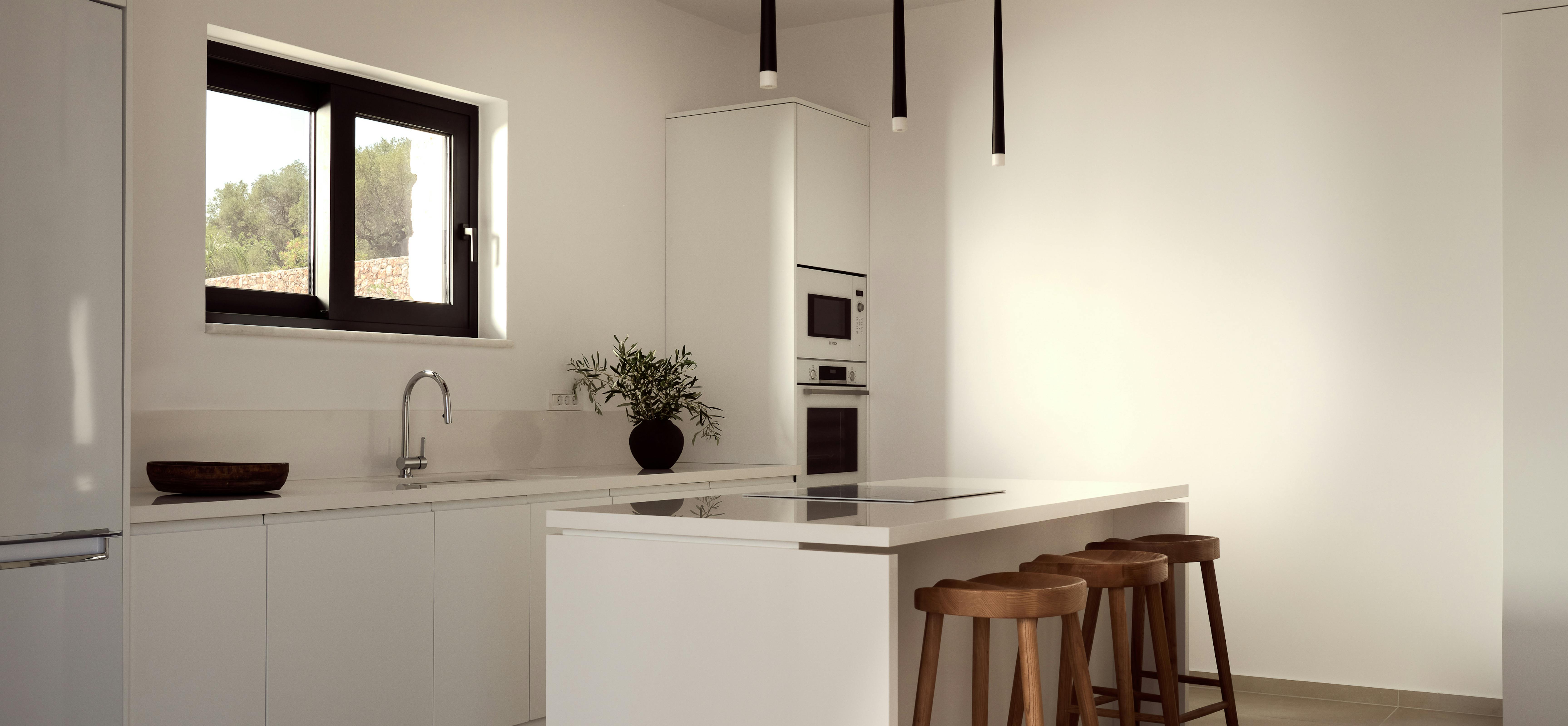 A contemporary white kitchen with sleek handleless cabinets, white waterfall-edge island with three wooden bar stools, black pendant lights, and recessed wooden ceiling detail.