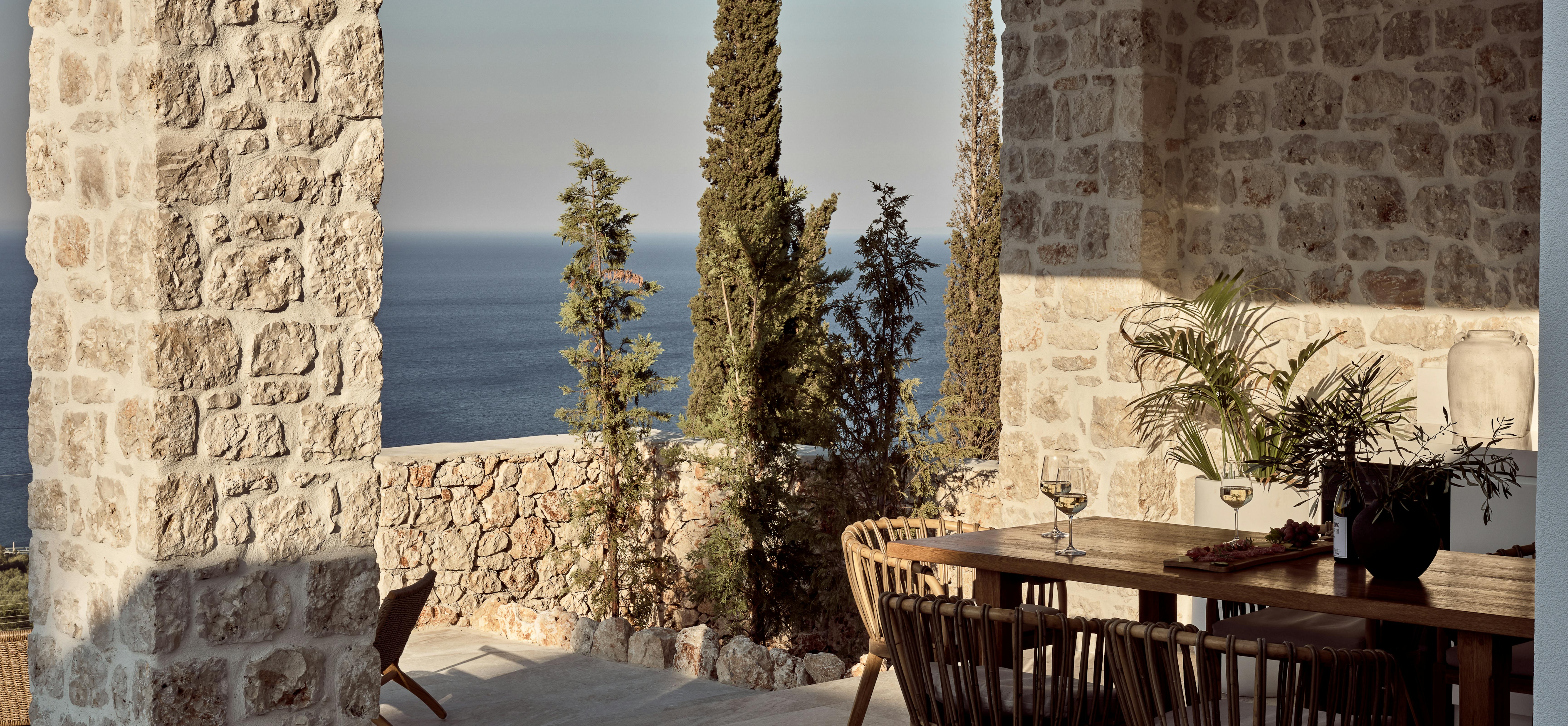 A covered outdoor dining terrace with rough stone columns framing views of cypress trees and the Mediterranean Sea, featuring a wooden dining table with woven chairs and modern lounge seating.