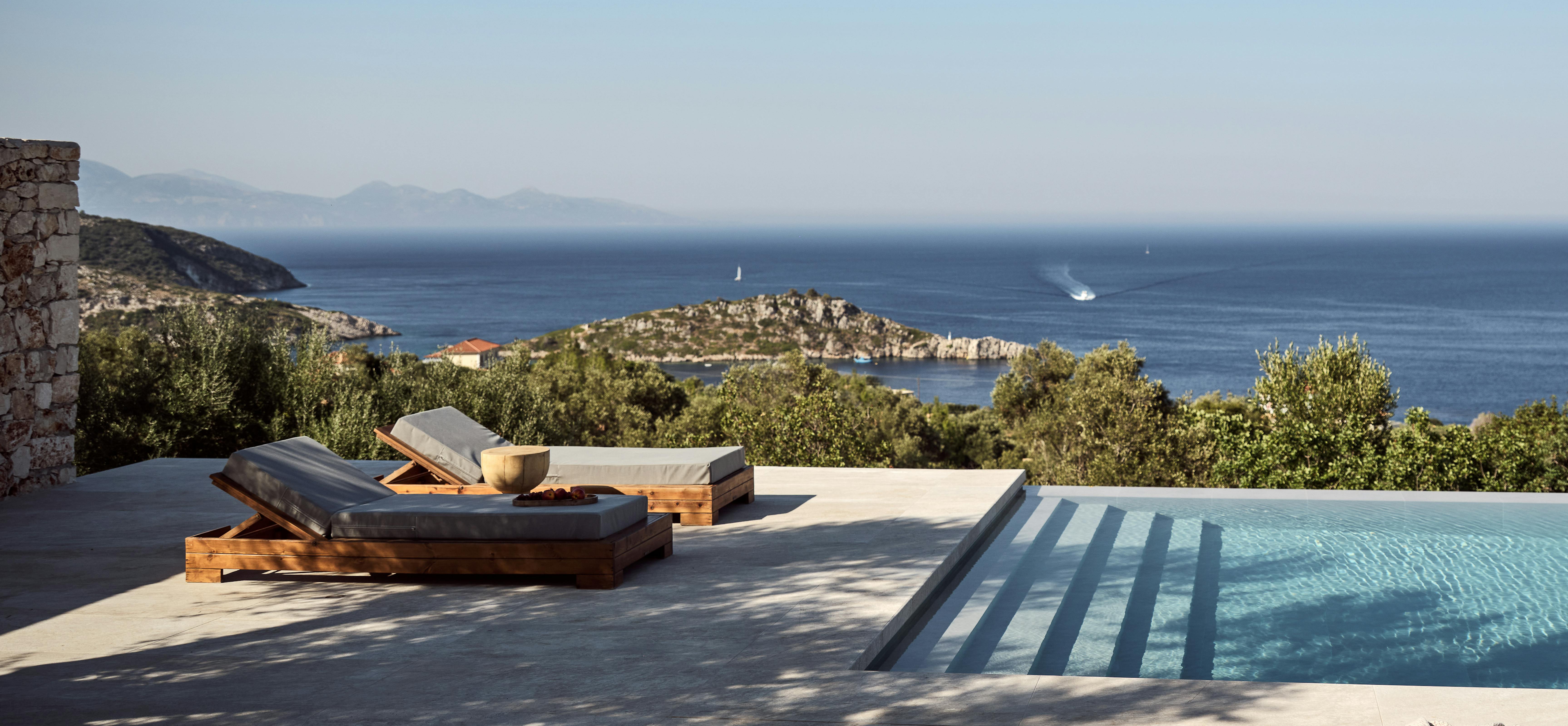 A minimalist infinity pool terrace with two wooden sun loungers, panoramic views of the deep blue Mediterranean Sea, rocky coastal islands, sailboats, and mountains in the hazy distance.