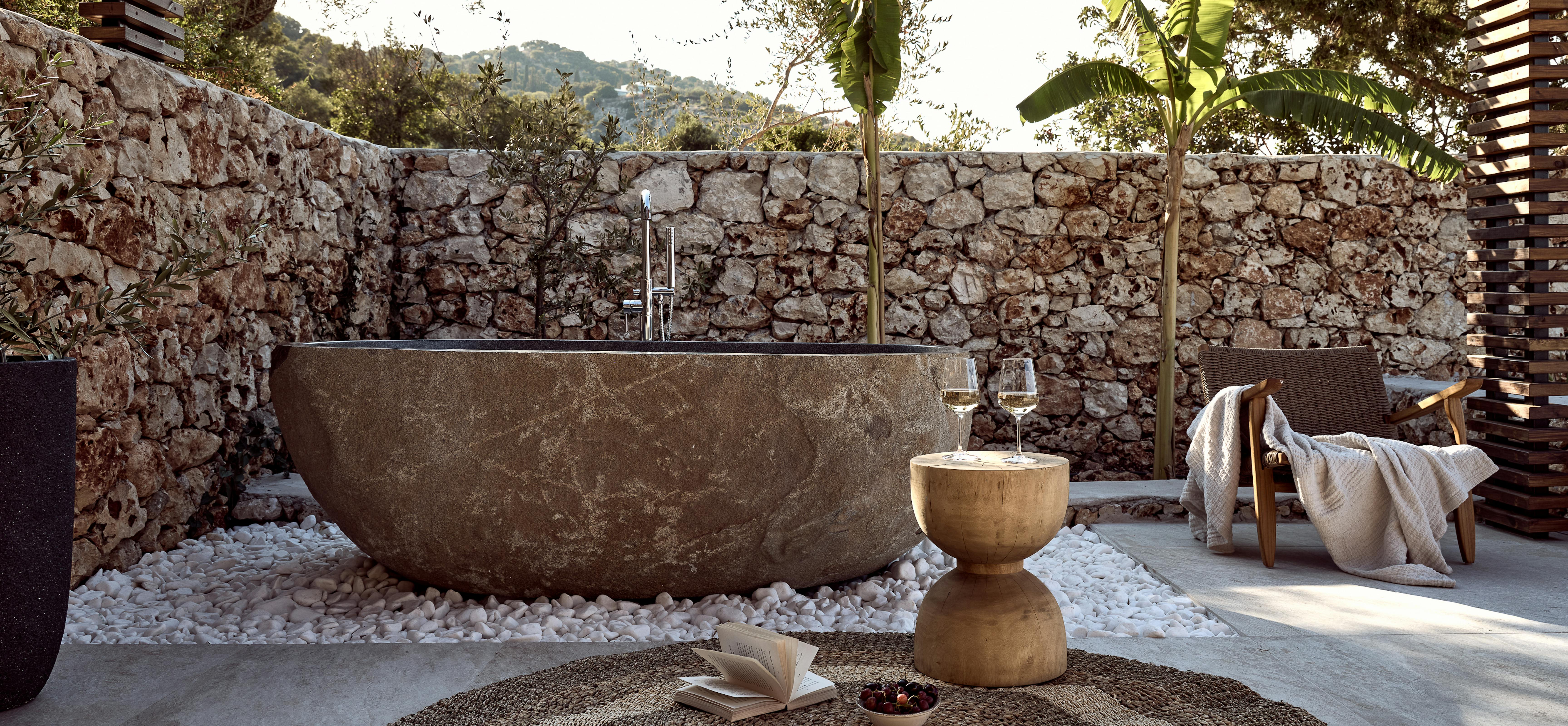 A luxurious outdoor bathing area with a large stone soaking tub on white pebbles, surrounded by dry-stacked stone walls, banana plants, olive trees, wooden slatted screens, and a woven lounge chair with throws, featuring a sculptural wooden side table with wine glasses.