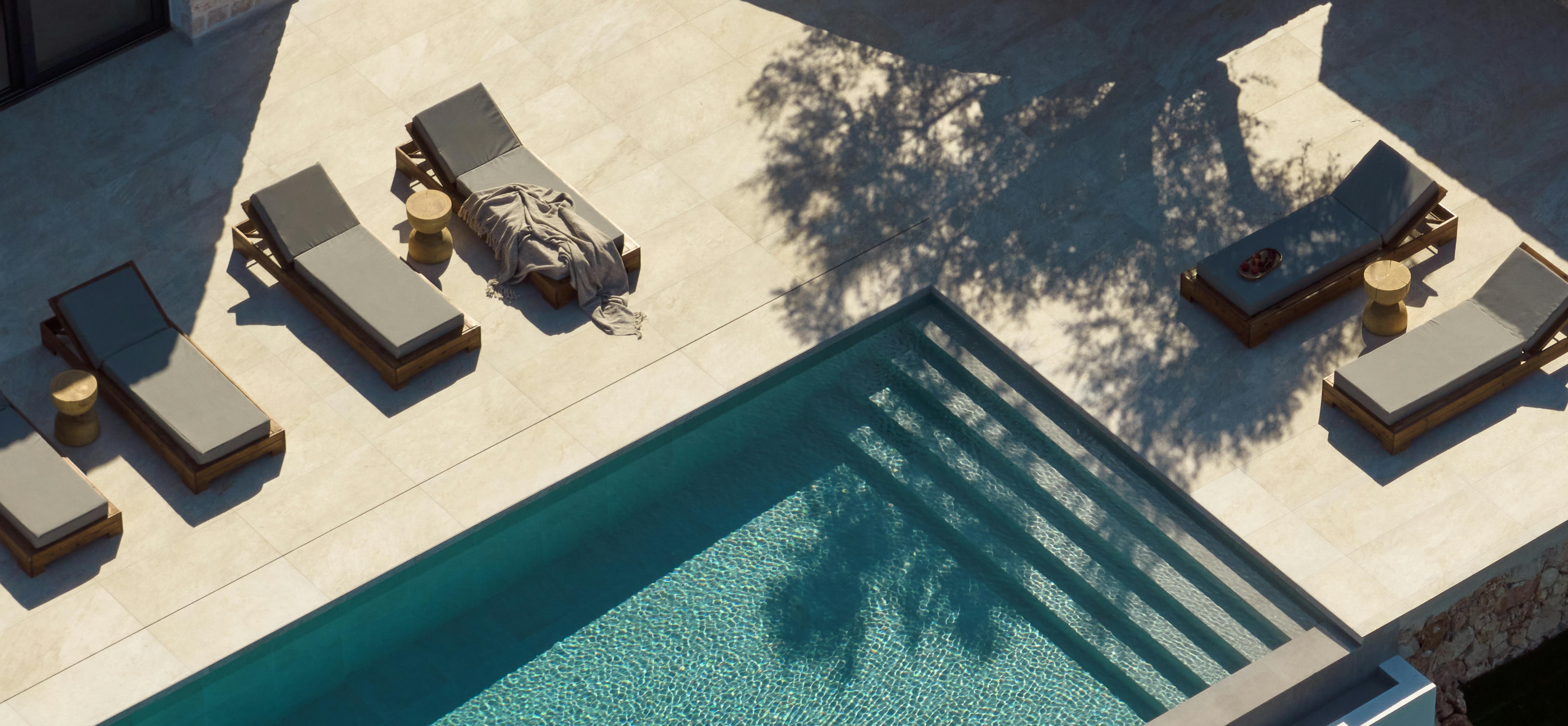 An aerial view of a modern rectangular swimming pool with turquoise water and built-in steps, surrounded by pale stone decking with wooden sun loungers topped with grey cushions, brass side tables, and curved stone retaining walls, with dappled tree shadows across the deck.