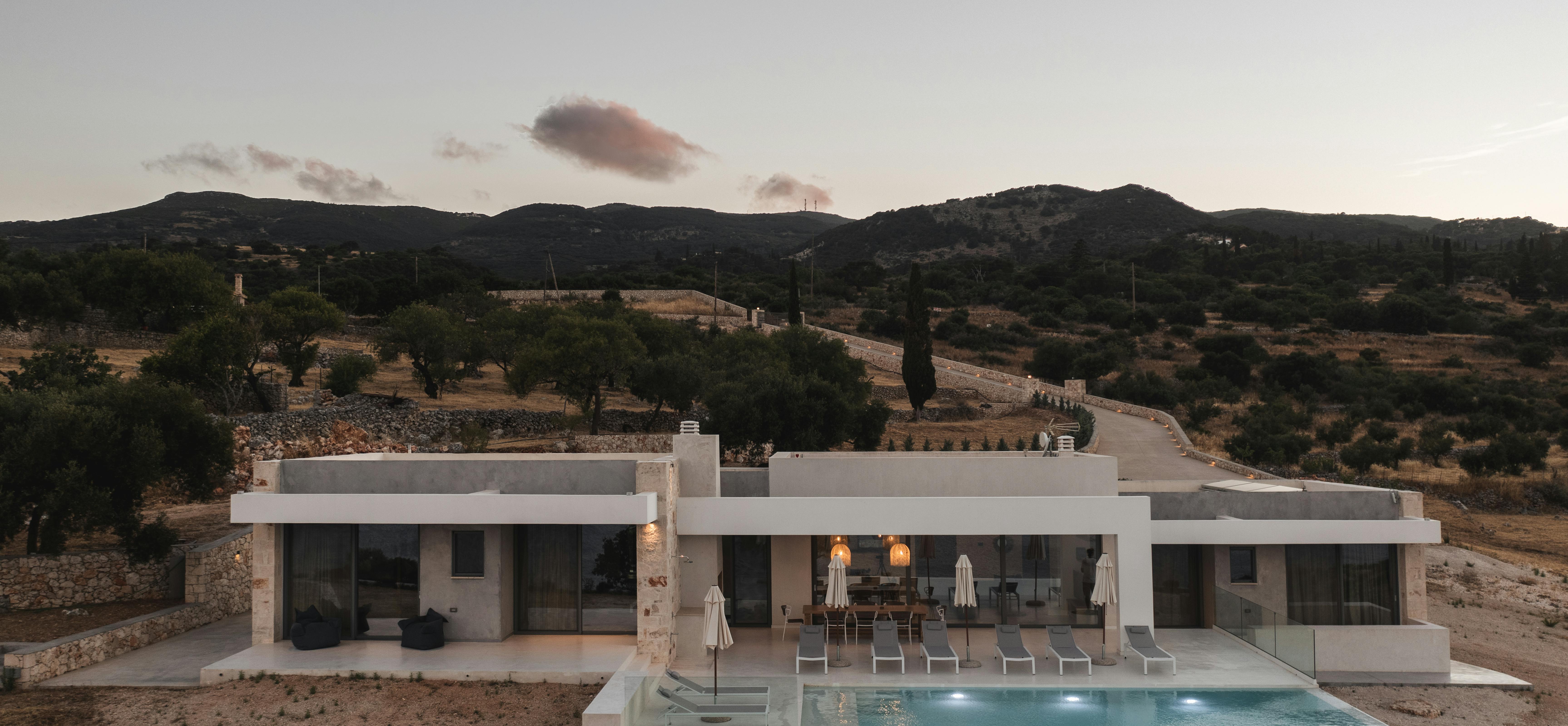 A single-story white villa with clean modernist lines and an illuminated infinity pool perches on a Mediterranean hillside at dusk, surrounded by natural terrain.
