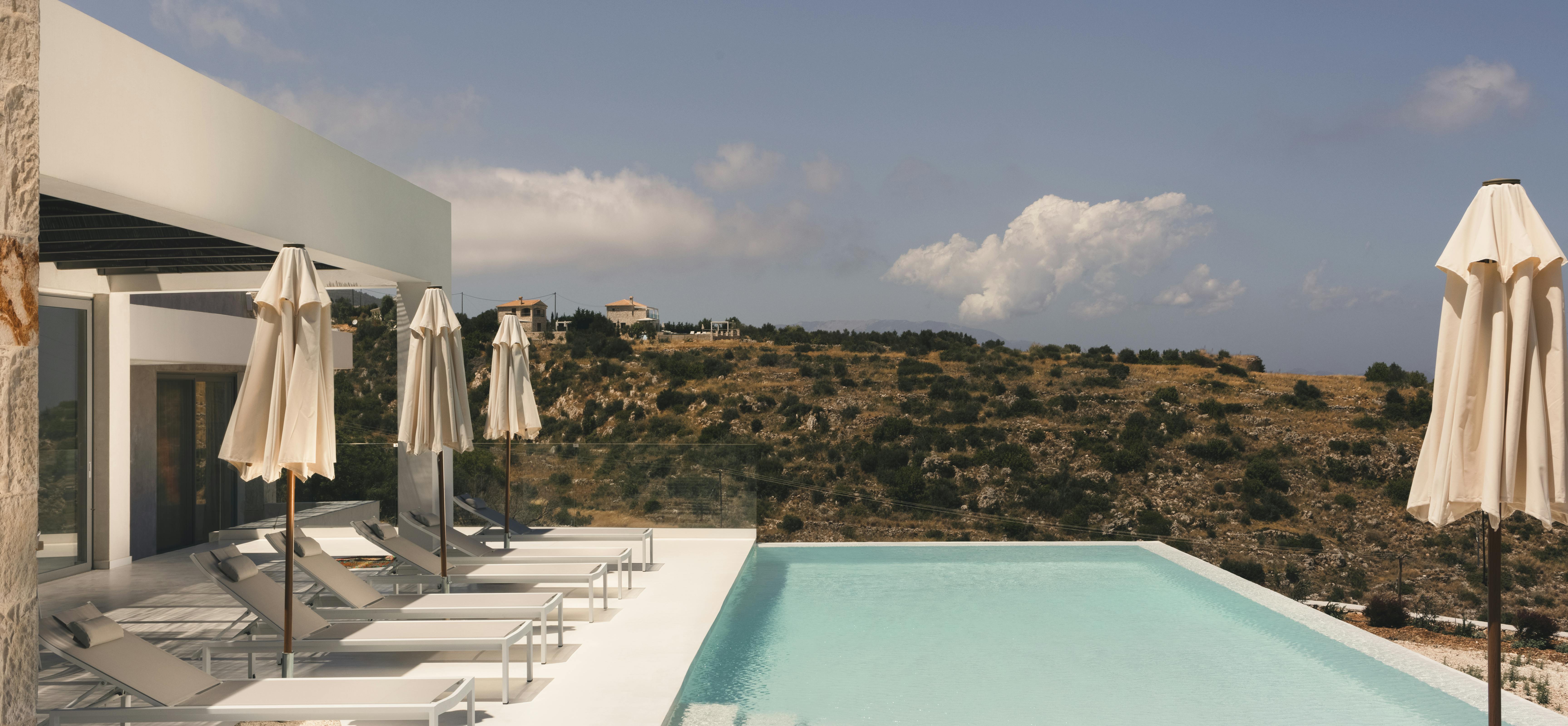 A modern white villa with a pristine infinity pool sits on a hillside, offering panoramic views of the Mediterranean landscape and sea under a bright blue sky with scattered clouds.