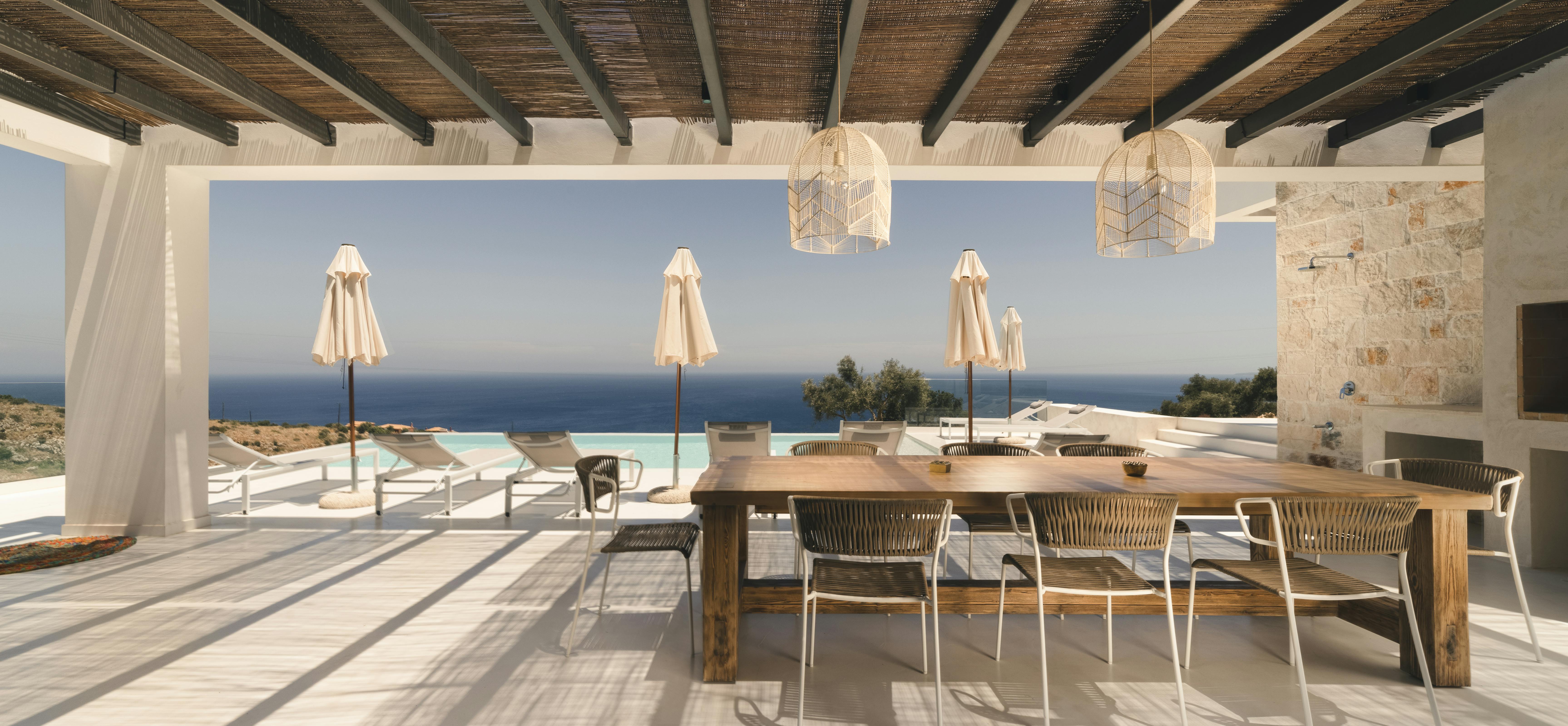 A covered outdoor terrace with traditional reed ceiling features a rustic wooden dining table, woven pendant lights, and dramatic views of the infinity pool and sea horizon.