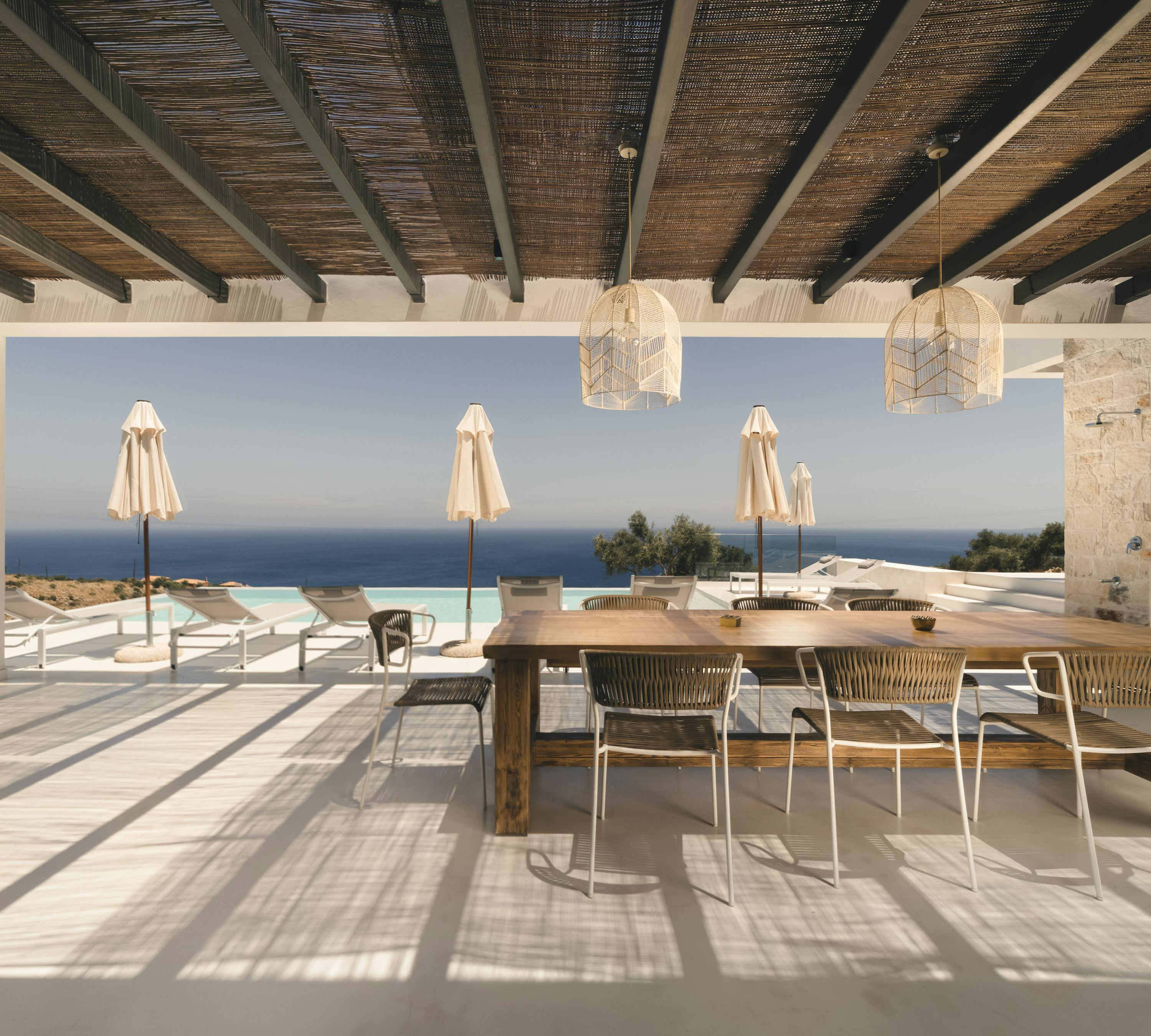 A covered outdoor terrace with traditional reed ceiling features a rustic wooden dining table, woven pendant lights, and dramatic views of the infinity pool and sea horizon.