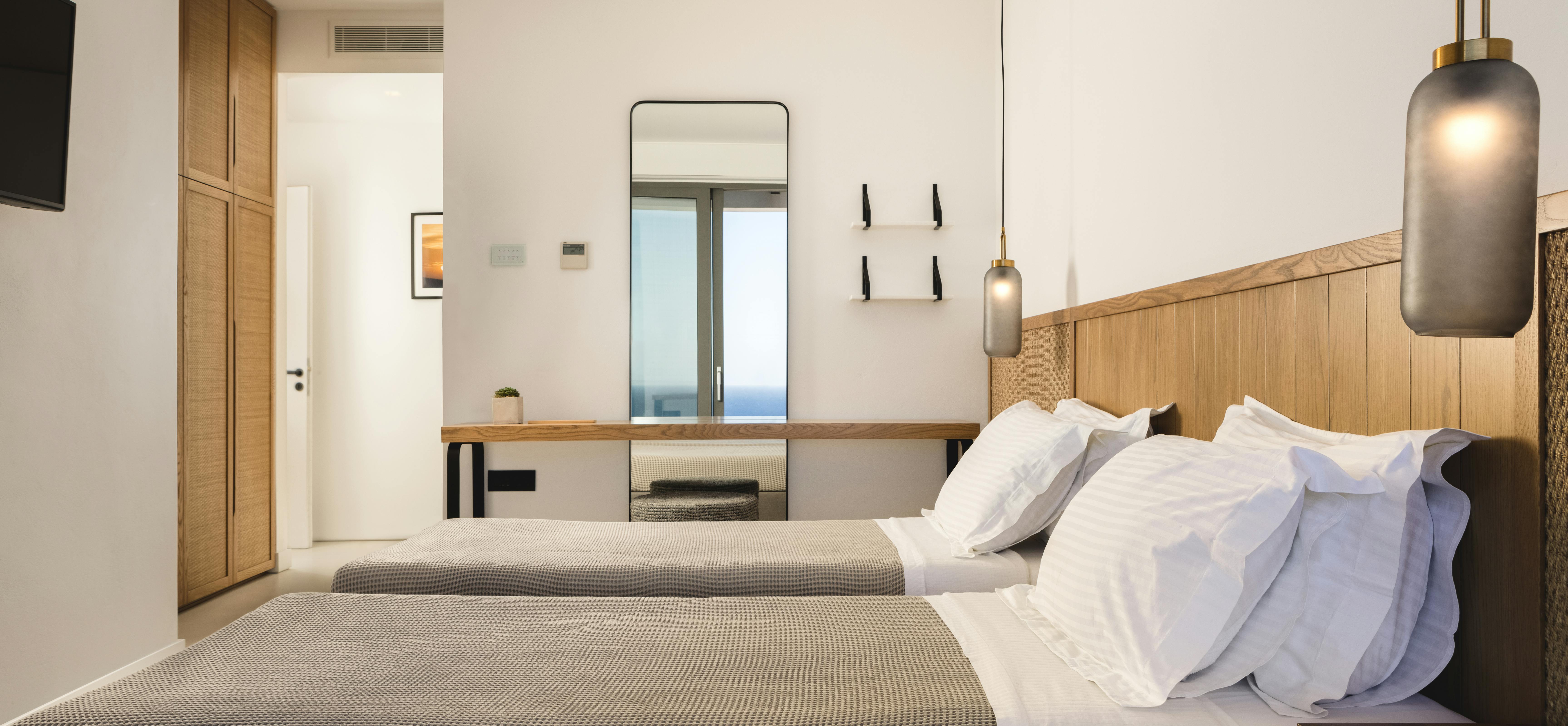 A twin bedroom with two beds features oak headboards, textured bedding, modern pendant lights with brass details, and a window framing distant sea views.