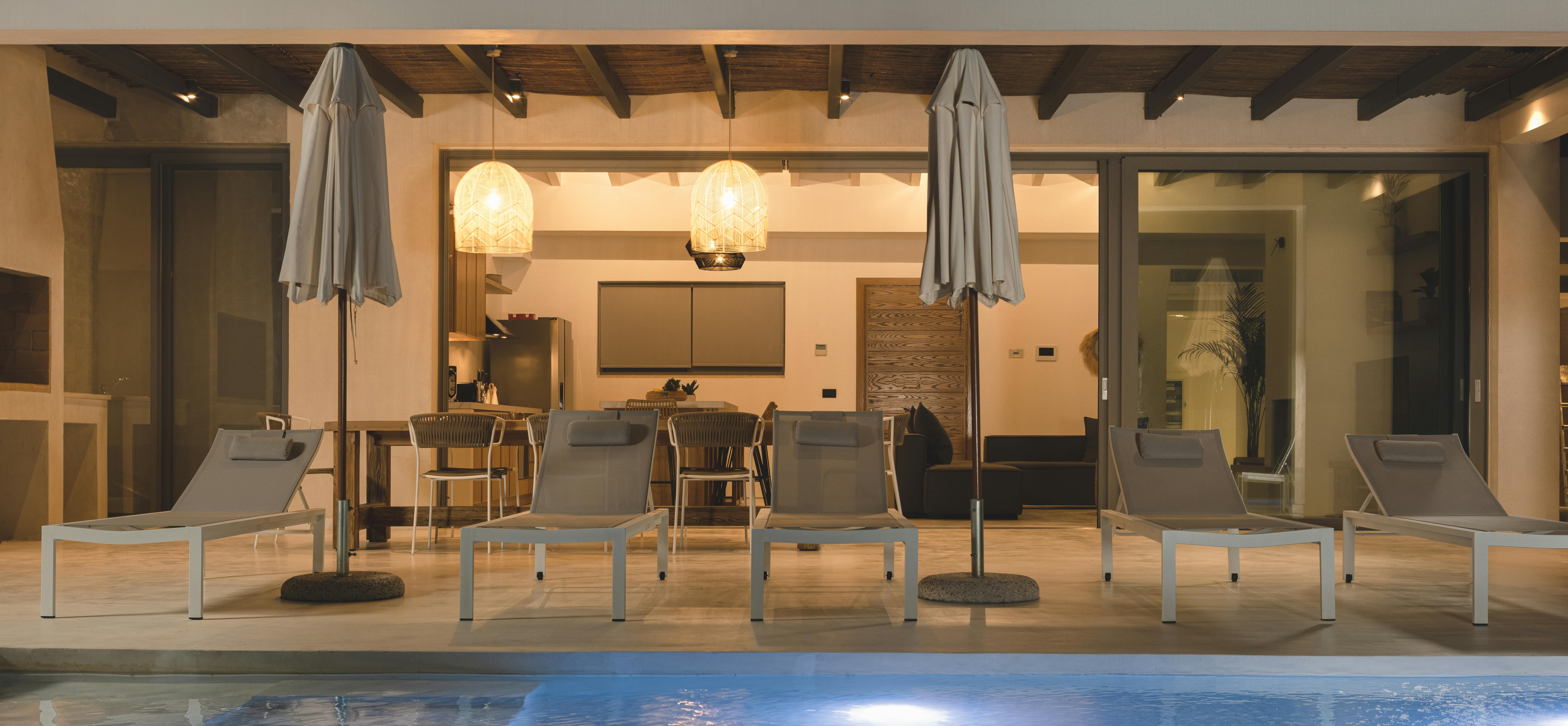 A nighttime view of the pool terrace features a row of white modern loungers with furled umbrellas positioned along the illuminated infinity pool, with the warmly lit interior visible through floor-to-ceiling glass doors and exposed beam ceiling.