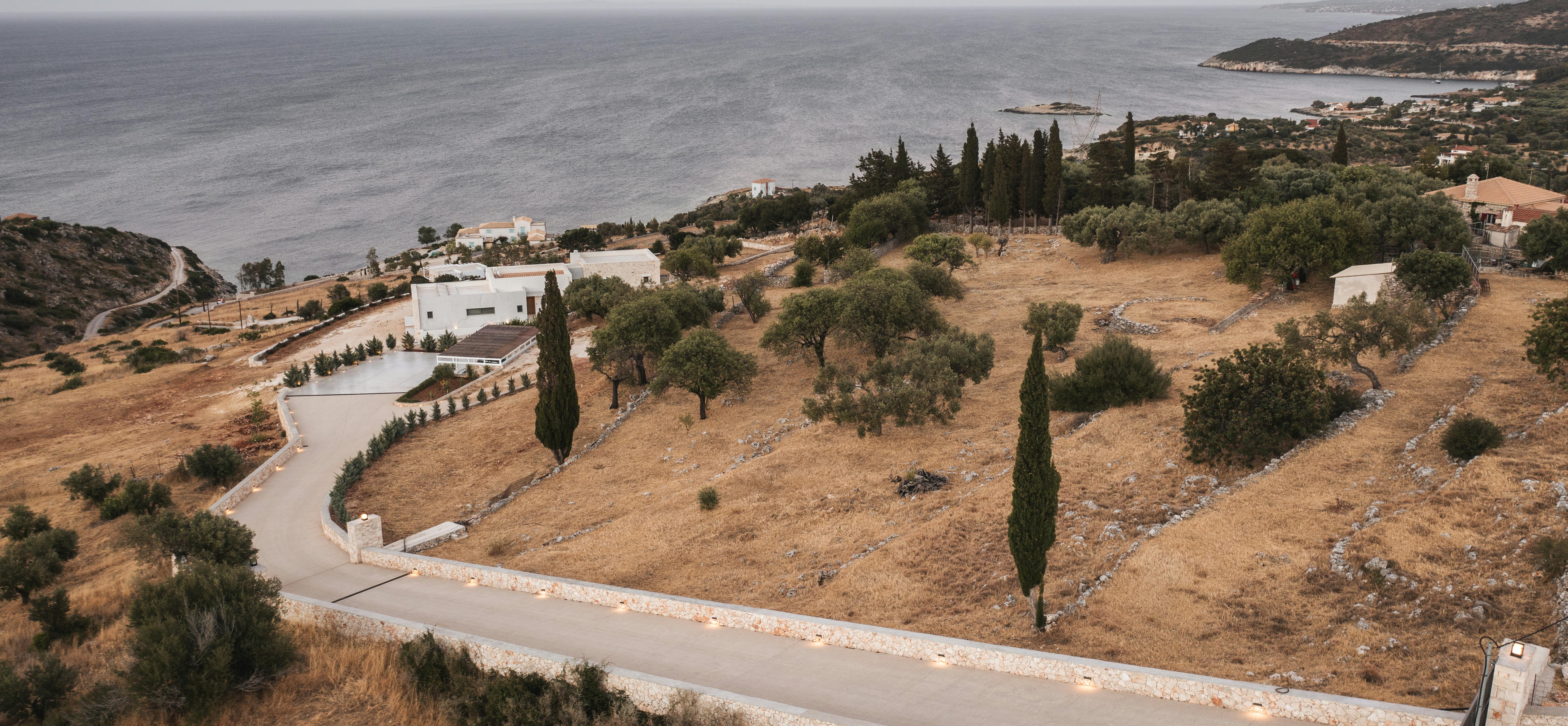The infinity pool glows with underwater lighting at night, its still surface reflecting the warmly illuminated villa interior with woven pendant lights, while white loungers and closed umbrellas line the deck under the dark evening sky.