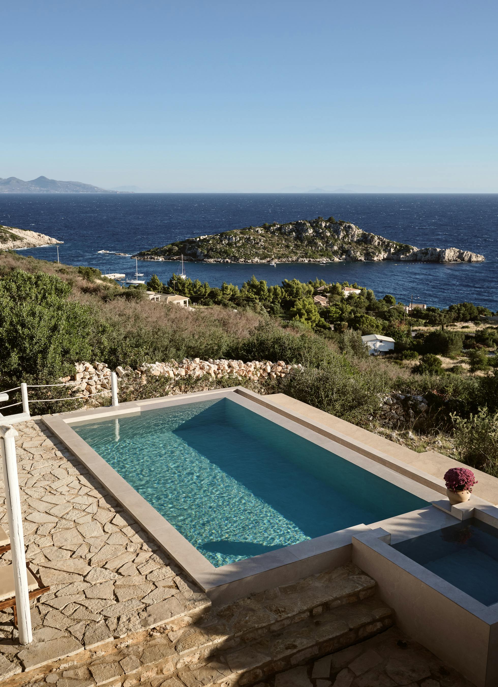 A multi-level infinity pool complex built into the hillside features stone terracing, white pergolas providing shade over wooden loungers, and sweeping views across a protected bay with rocky islands to the mountainous coastline beyond.