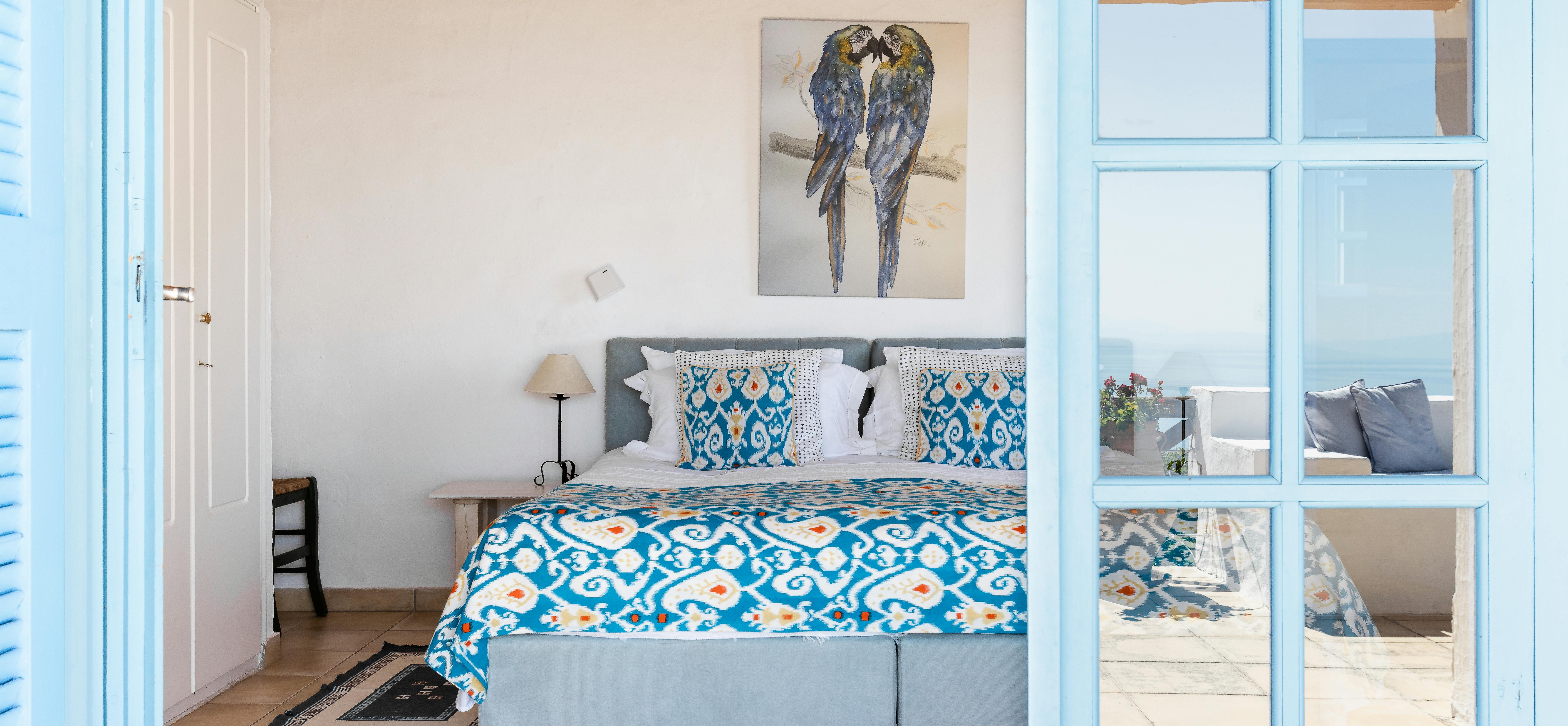 Bright blue French doors with multiple glass panes frame a view into a whitewashed bedroom with twin beds covered in colourful ikat throws and decorative pillows, exposed ceiling beams, artwork of perched birds, and glimpses of the pool terrace beyond.