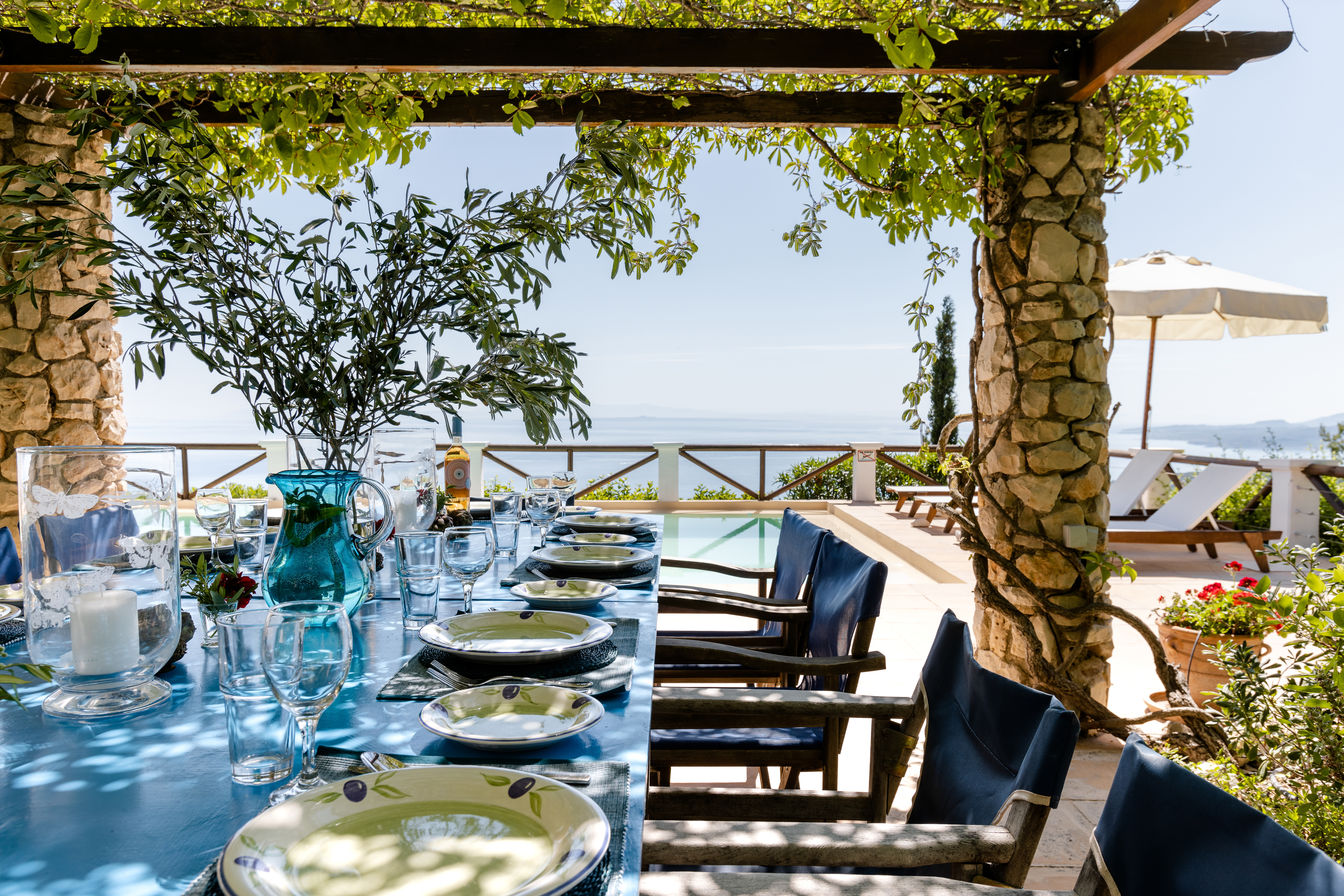 A vine-draped dining pergola frames an inviting outdoor table covered in bright blue cloth and set with olive-patterned dishes and crystal stemware, with blue director's chairs arranged along both sides overlooking an infinity pool and the shimmering sea beyond under dappled sunlight.