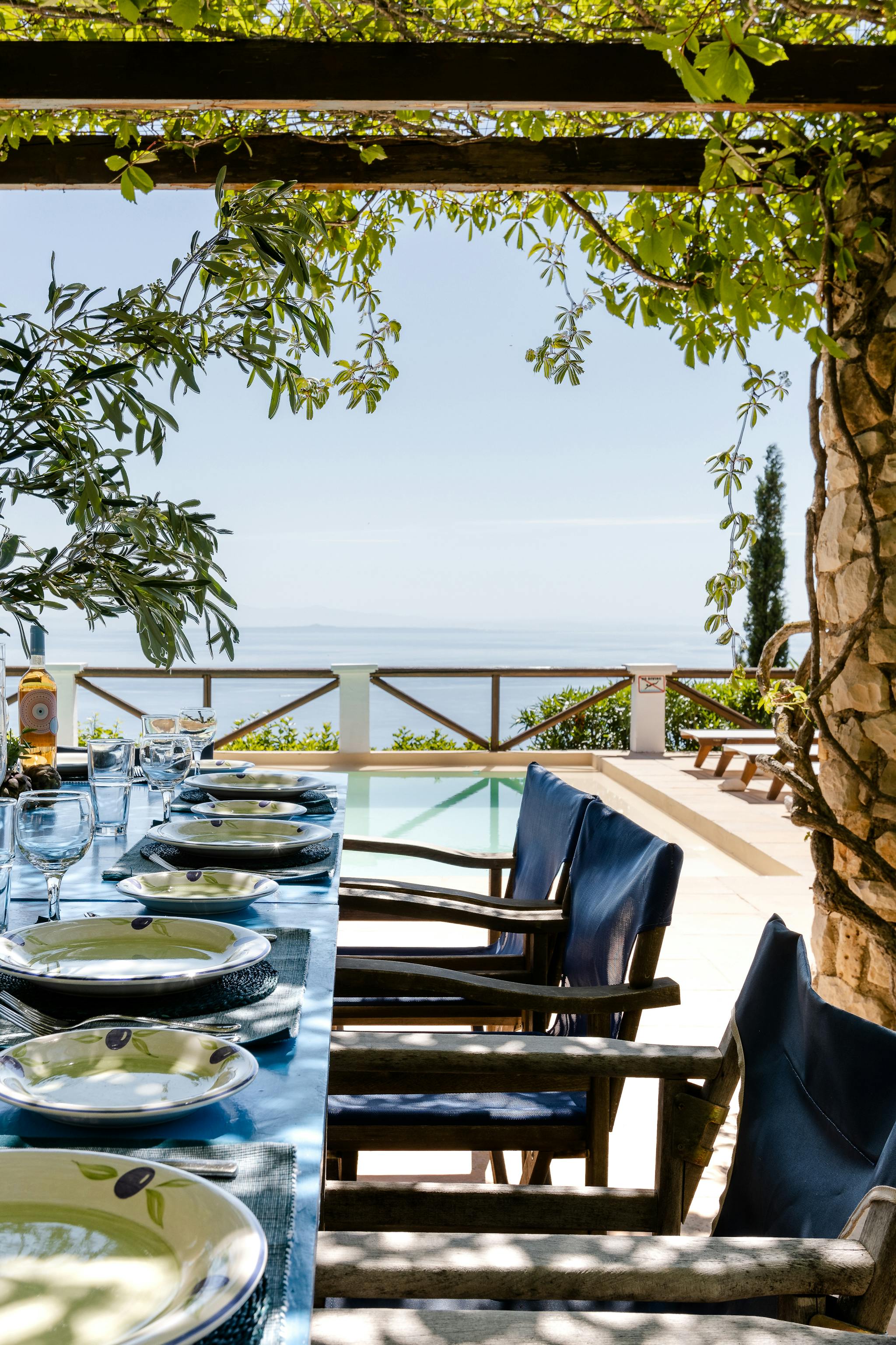 A vine-draped dining pergola frames an inviting outdoor table covered in bright blue cloth and set with olive-patterned dishes and crystal stemware, with blue director's chairs arranged along both sides overlooking an infinity pool and the shimmering sea beyond under dappled sunlight.