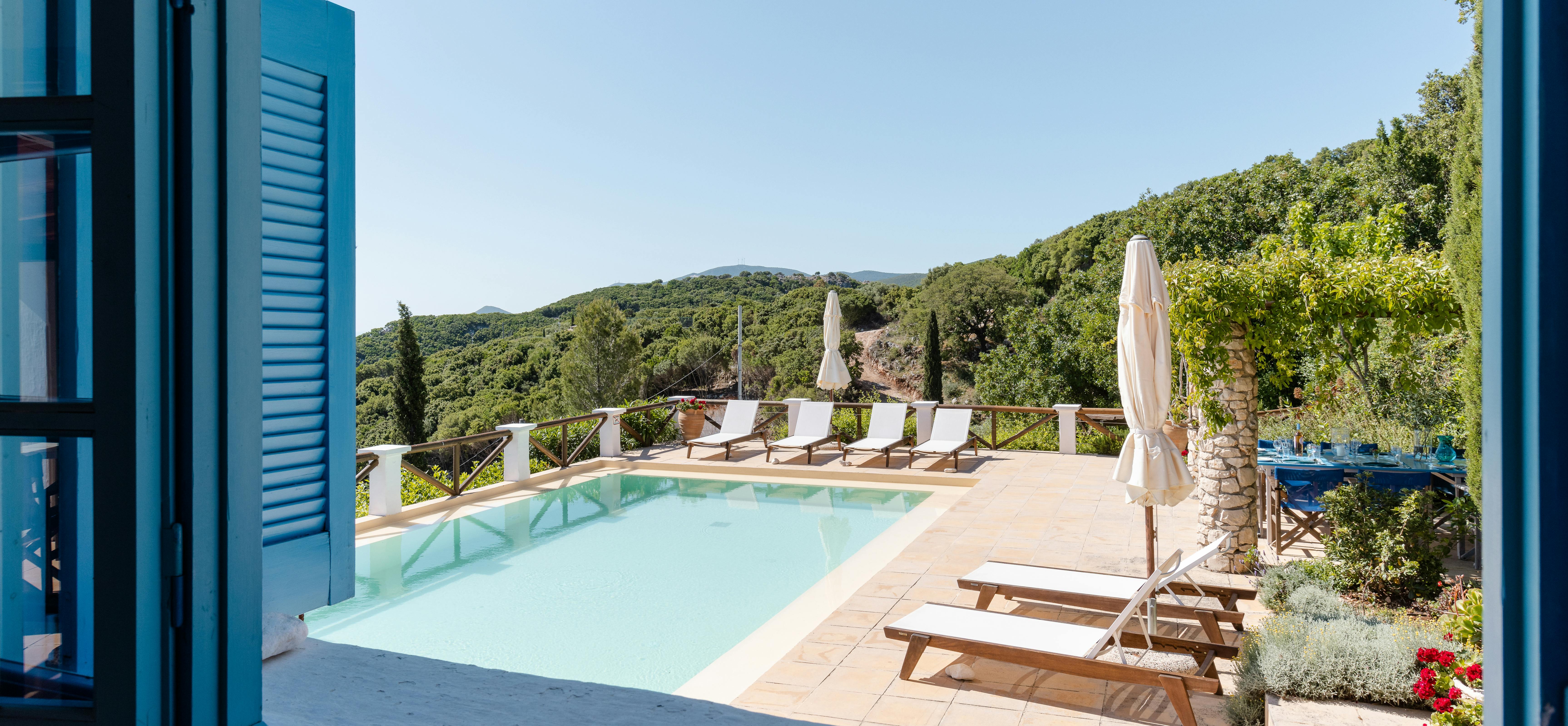 A stunning view through blue-painted louvered shutters and French doors reveals the infinity pool terrace with cream umbrellas, wooden sun loungers, white safety railings, and sweeping vistas across forested hillsides to the distant sea under endless blue skies.