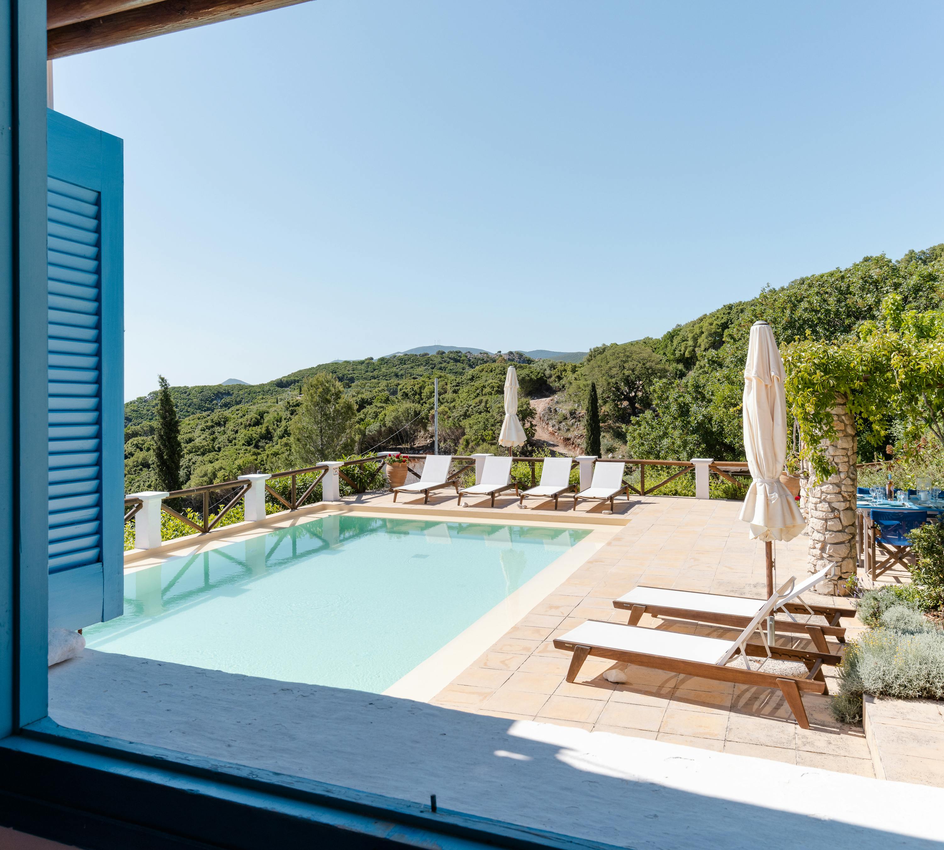 A stunning view through blue-painted louvered shutters and French doors reveals the infinity pool terrace with cream umbrellas, wooden sun loungers, white safety railings, and sweeping vistas across forested hillsides to the distant sea under endless blue skies.