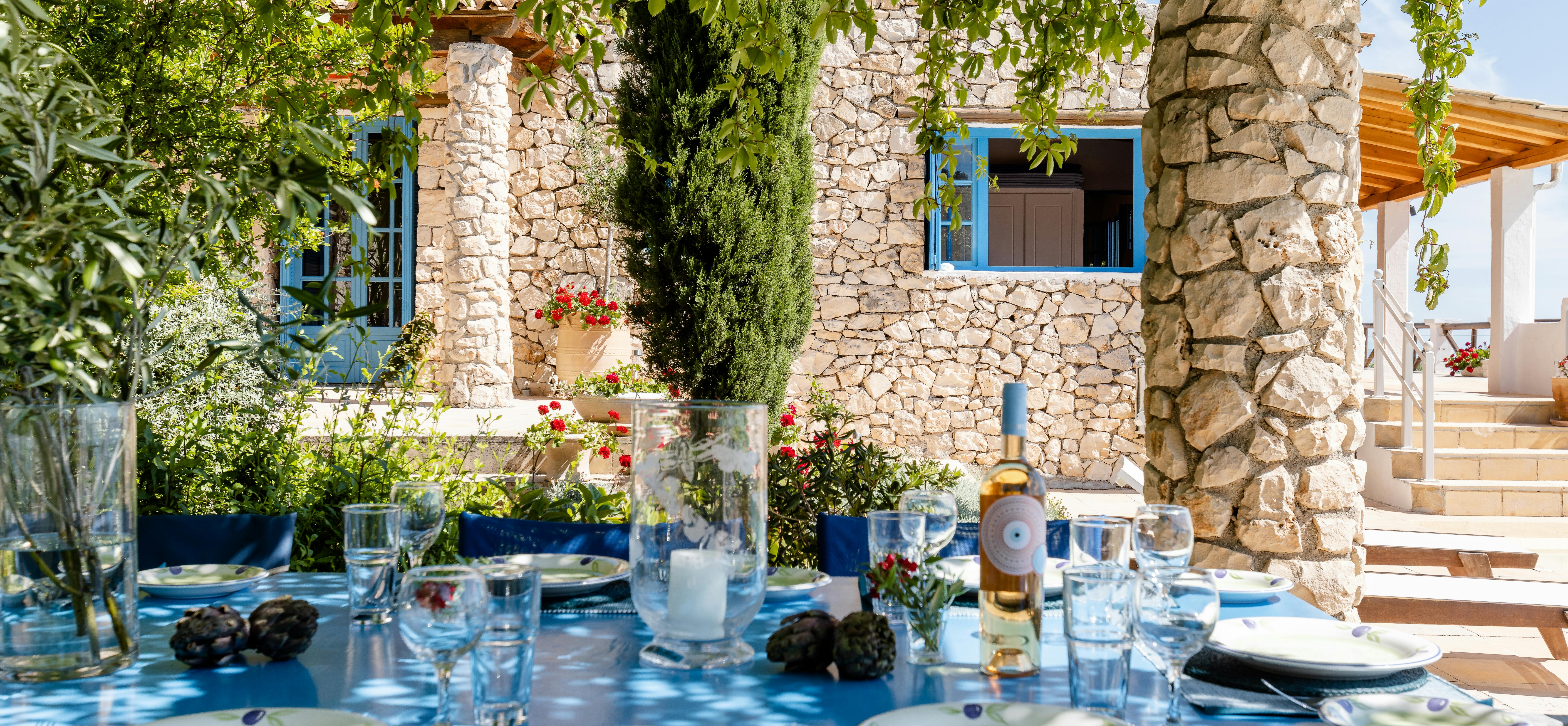 An elegantly set outdoor dining table draped in vibrant blue tablecloth displays olive branch patterned plates, crystal glassware, candles, and fresh flowers under a rustic pergola beside a traditional stone villa with blue-trimmed windows overlooking lush Mediterranean gardens.