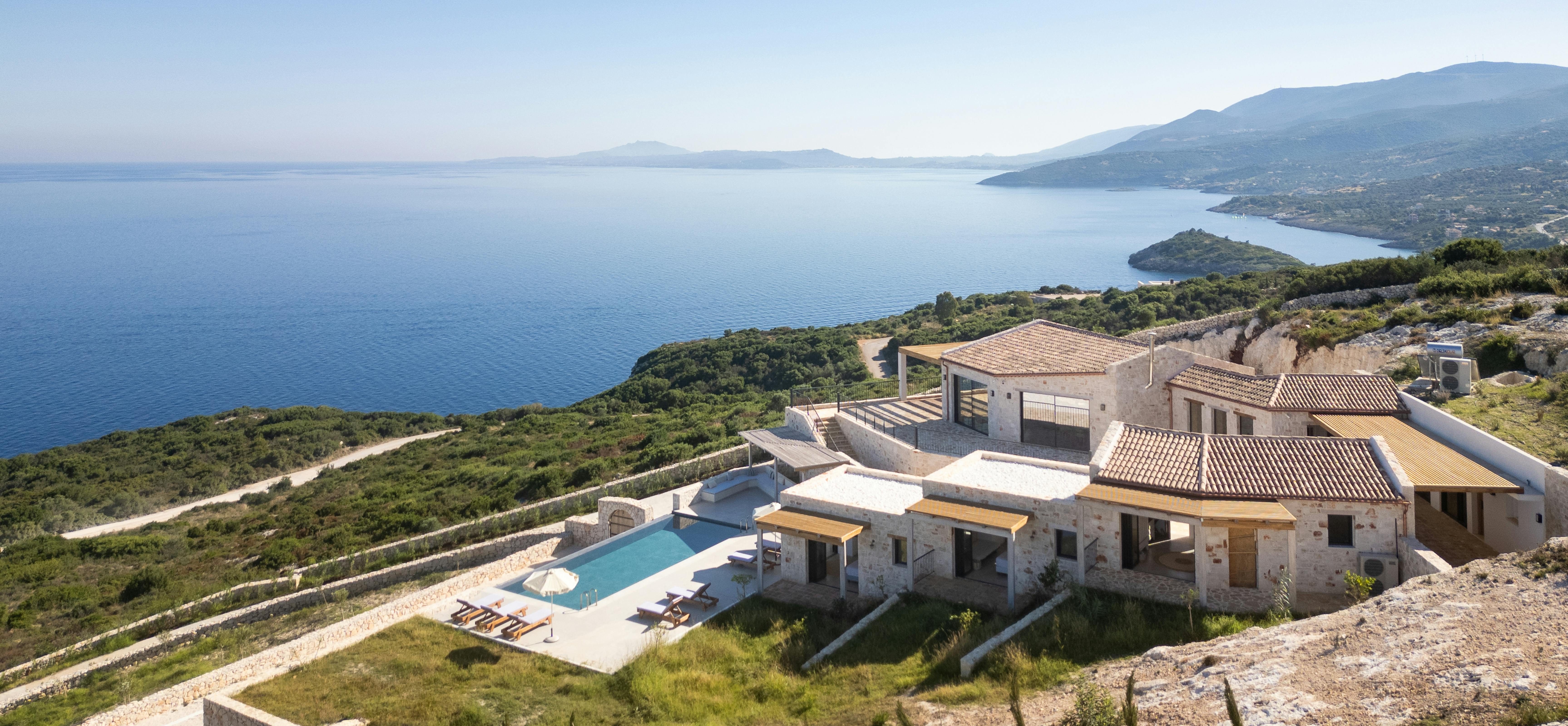 Aerial view of stone villa complex with terracotta roofs, infinity pool, and terraced grounds overlooking expansive bay and mountainous coastline.