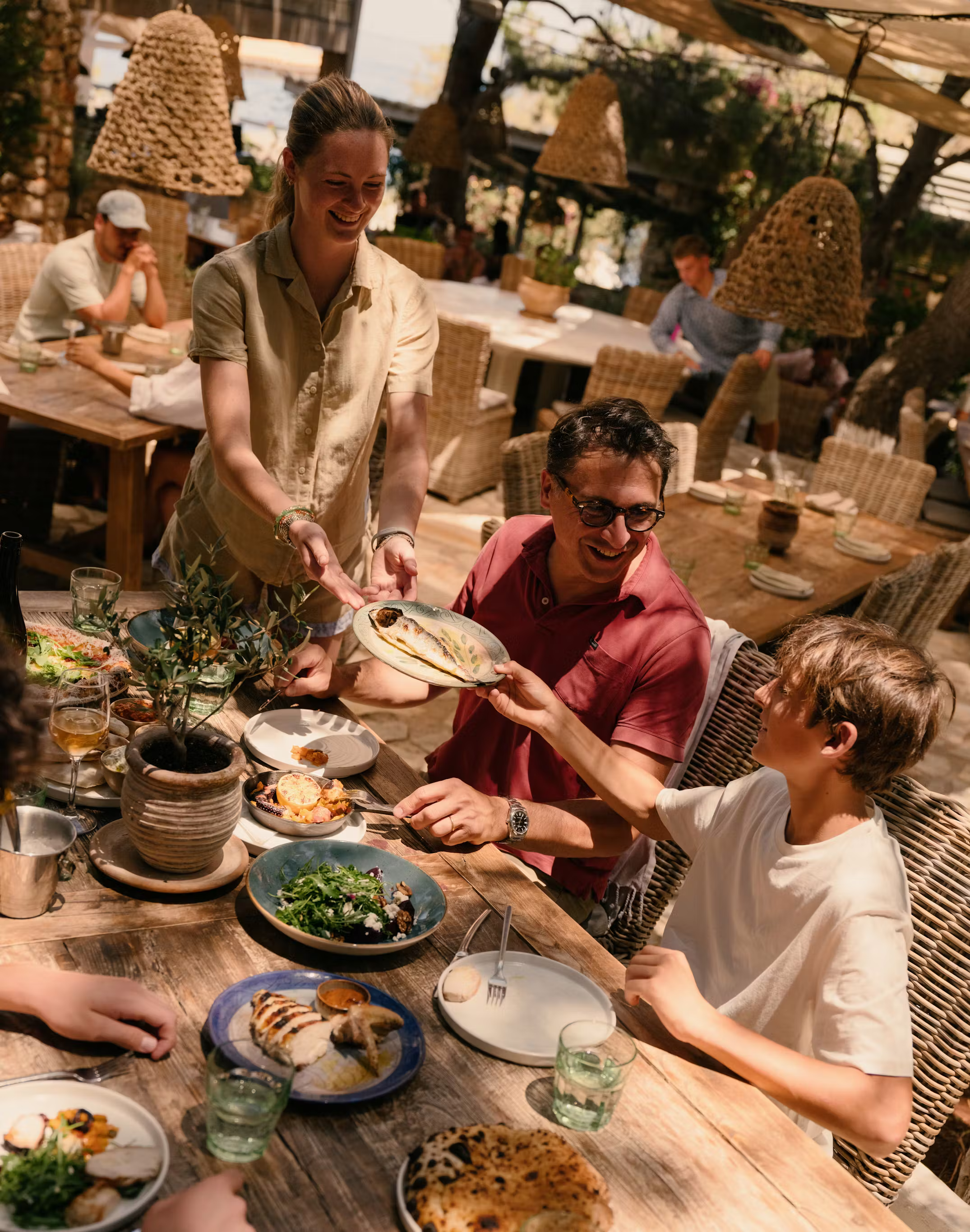 Server delivering plated fish dish to smiling family dining at rustic wooden table in Mediterranean restaurant with woven pendant lights.