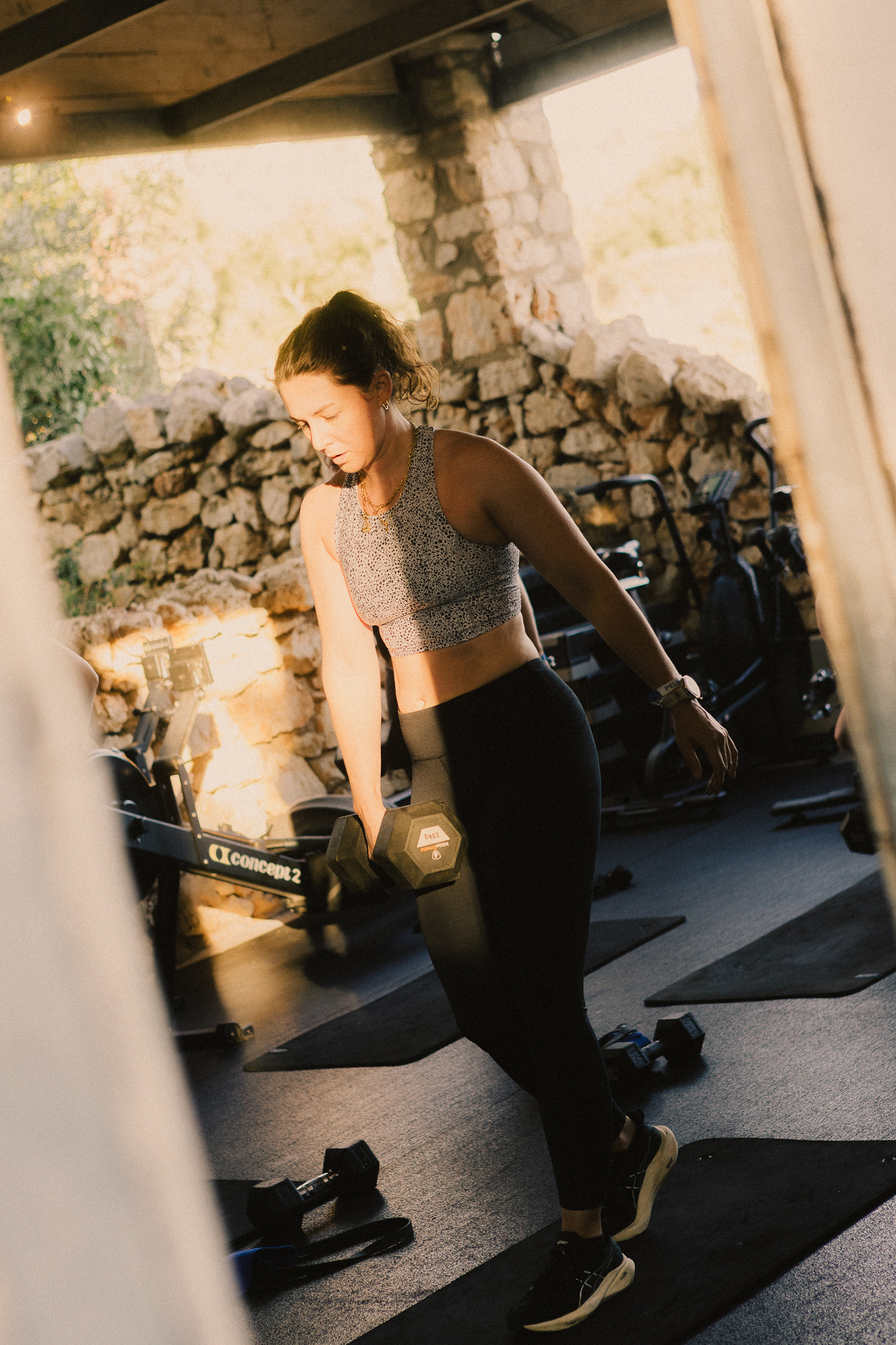 A woman in a grey patterned crop top and black leggings performs a lunge exercise with a dumbbell in a rustic gym with exposed stone walls and wooden beams. She's positioned on black exercise mats, with cardio equipment visible in the background. Natural light streams in, creating warm tones throughout the Mediterranean-style workout space.