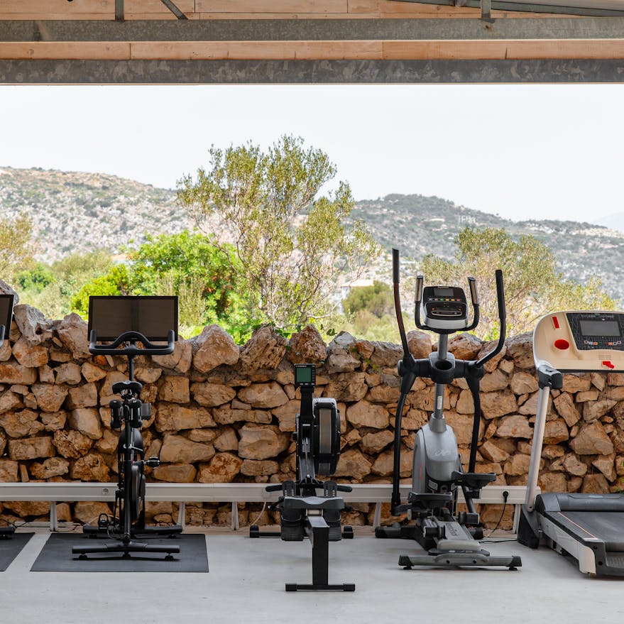An outdoor fitness area featuring a row of exercise equipment including two spin bikes, a rowing machine, and a treadmill arranged on black mats under a covered pavilion. The equipment faces a rustic stone wall backdrop with Mediterranean hillside views and olive trees visible beyond. Sheer beige curtains frame the sides of the open-air gym space, creating an elegant yet natural workout environment.